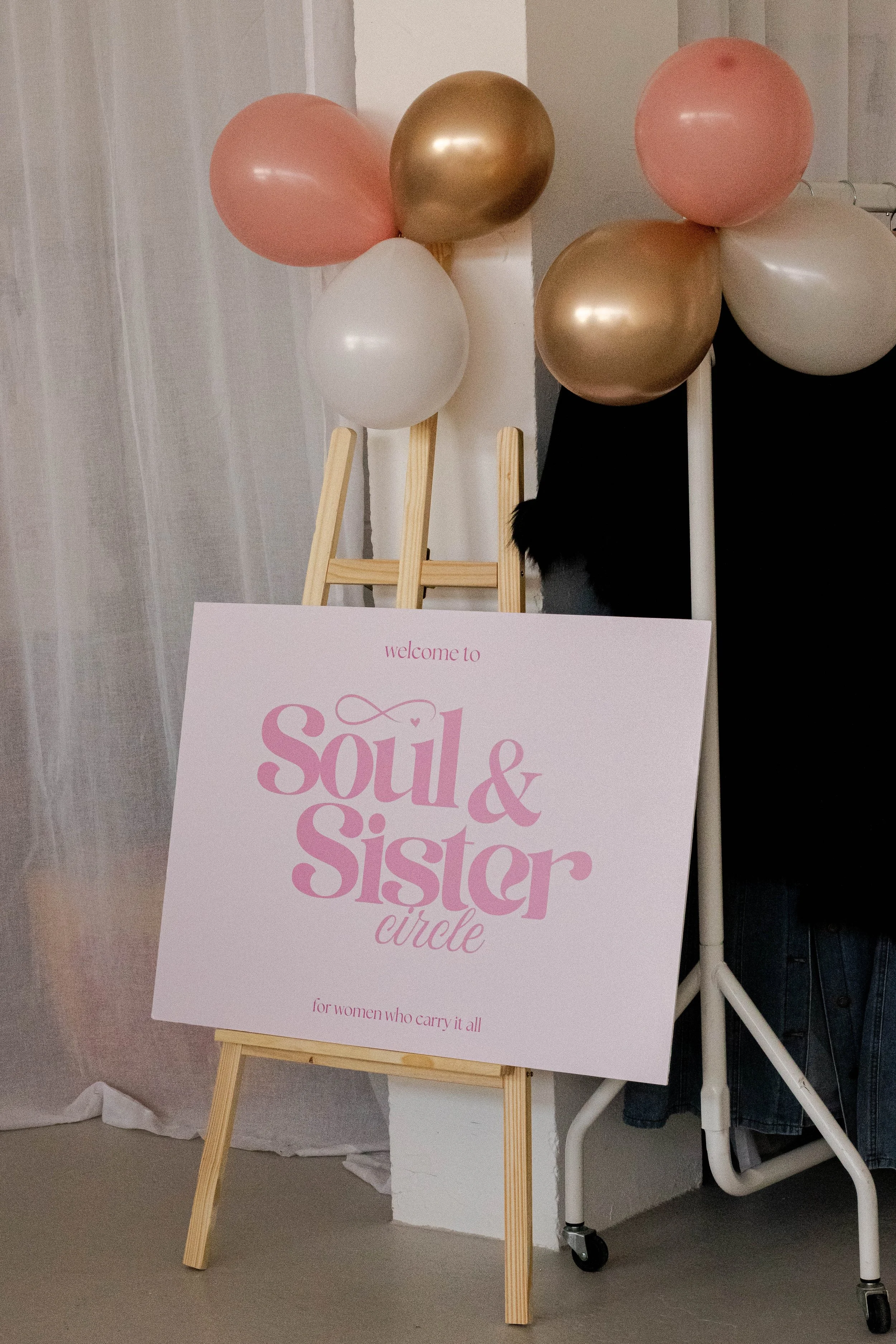 Welcome sign for 'Soul & Sister' circle with pink and gold balloons, a wooden easel, and a person partially visible behind the sign.