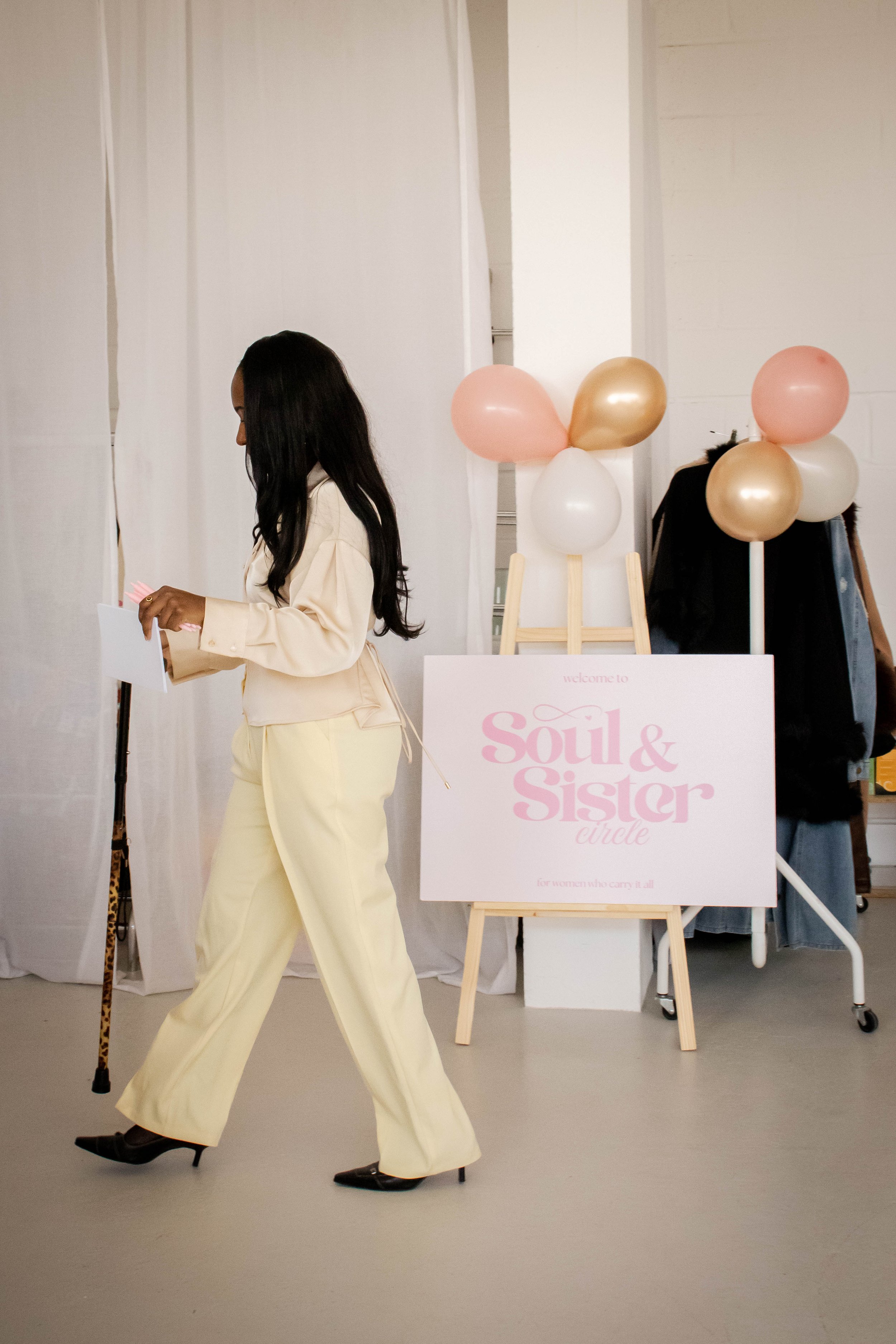 A woman walking past a sign that reads 'Soul & Sister Circle' with balloons and a clothing rack in the background.