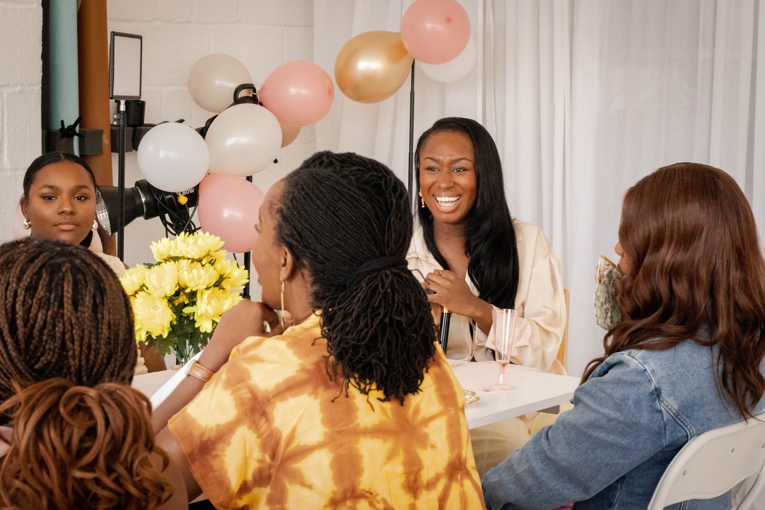 A group of women gathered around a table, celebrating with yellow flowers and balloons, with one woman speaking or laughing, in a decorated indoor setting.