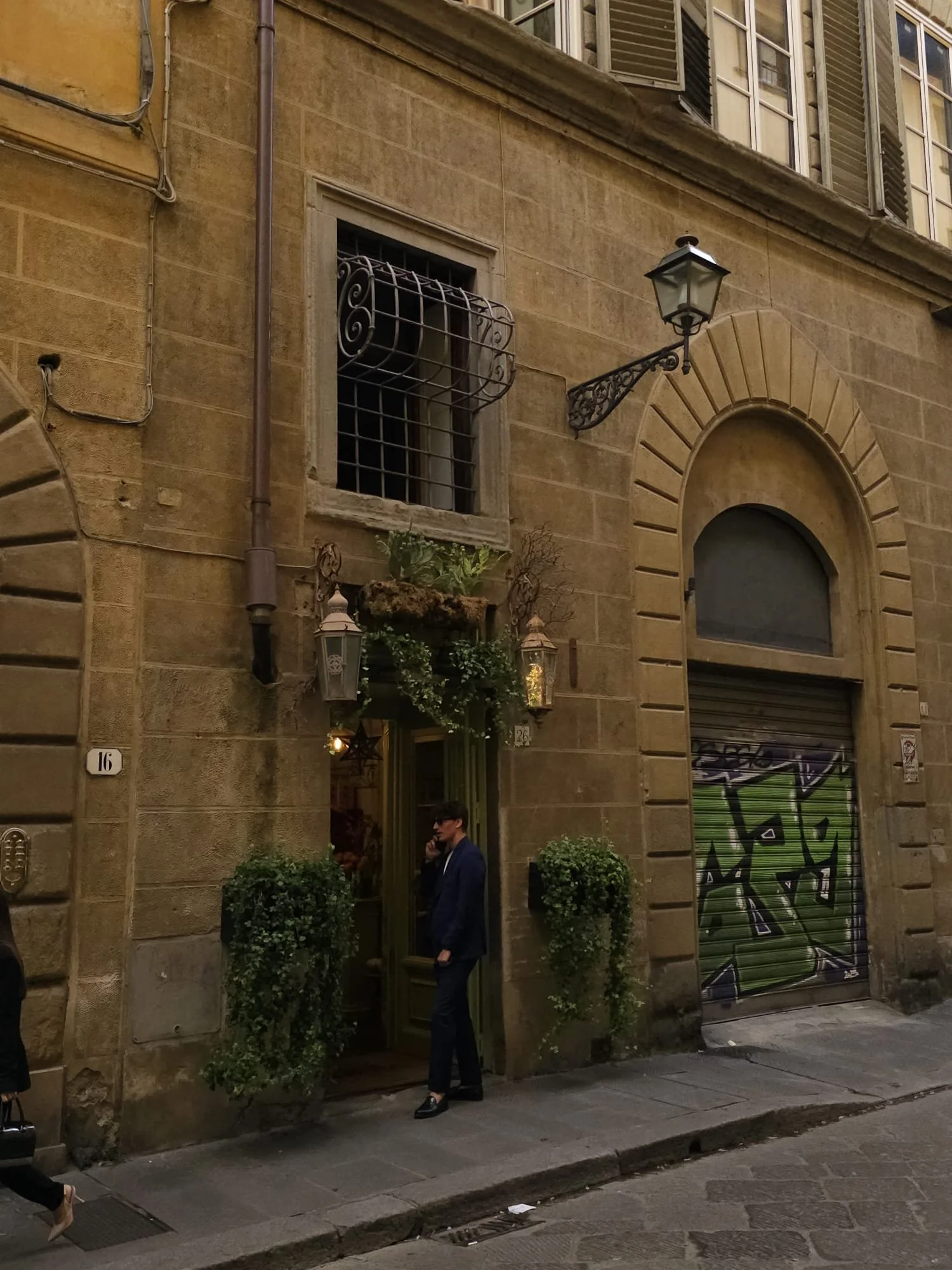 A man in a suit and sunglasses talking on a cell phone while standing outside a storefront on a cobblestone street, with an old building façade, a window with bars, decorative lanterns, and a graffiti-covered shutter in the background.