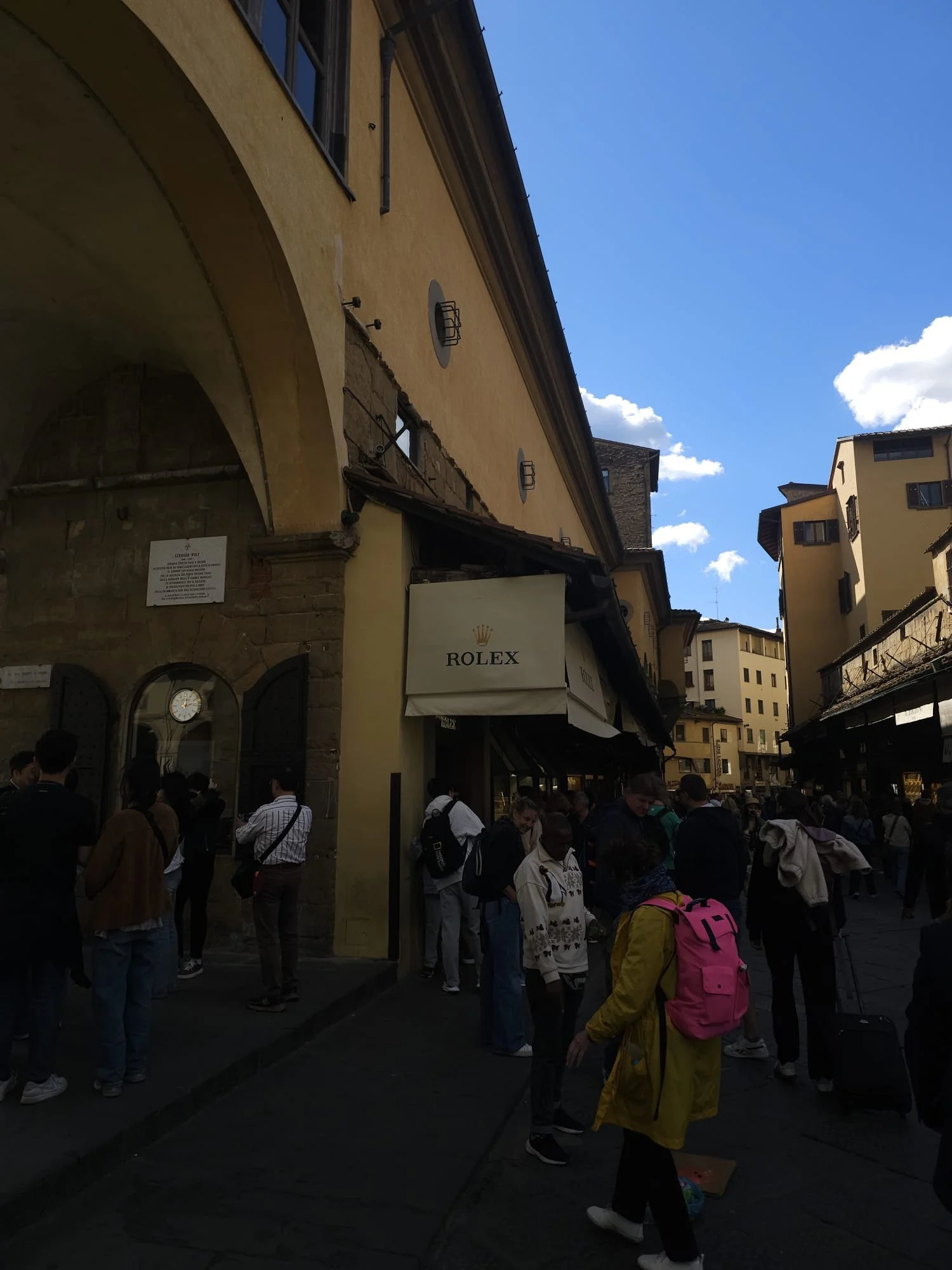 People standing outside a building with a Rolex sign, under a yellow wall with an archway. The scene appears to be in a busy street, with some individuals wearing backpacks and carrying luggage.