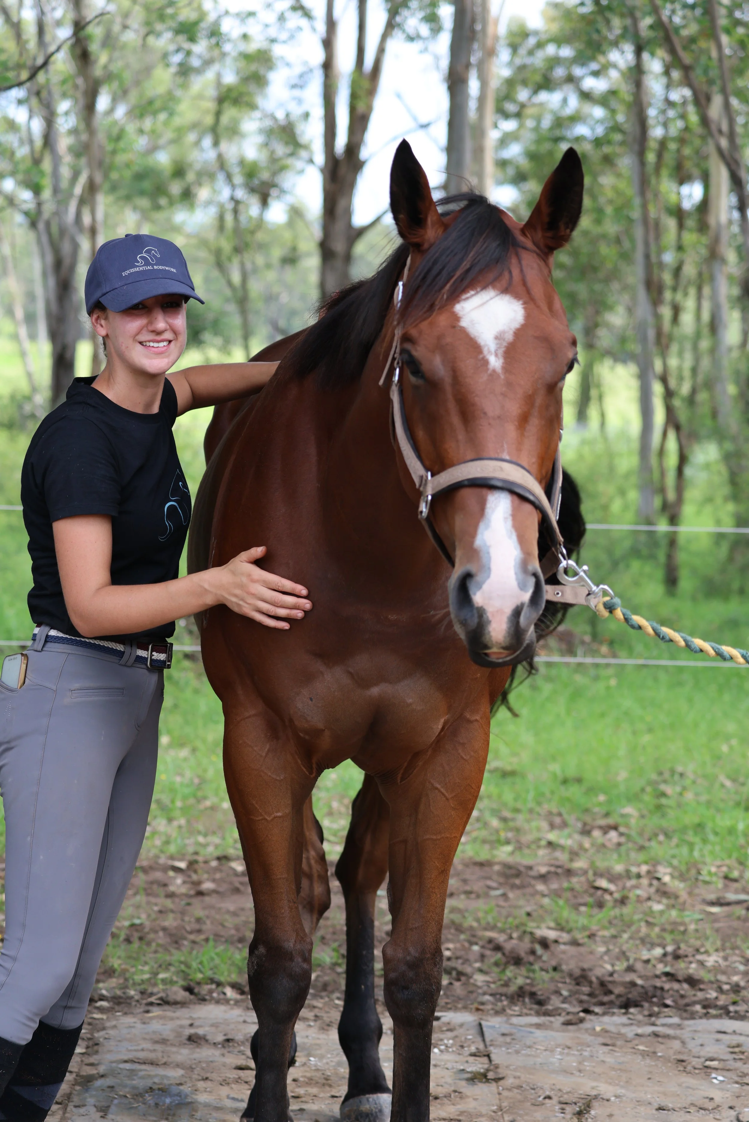 A woman petting a horse while a man works nearby. The woman is smiling and wearing a cap, gloves, and riding gear. The horse has a saddle and is standing calmly.