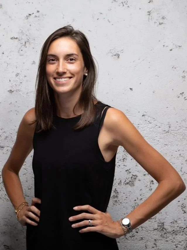 Una donna sorridente con capelli castani corti, indossa un top nero senza maniche e accessori, posa davanti a una parete bianca ruvida.