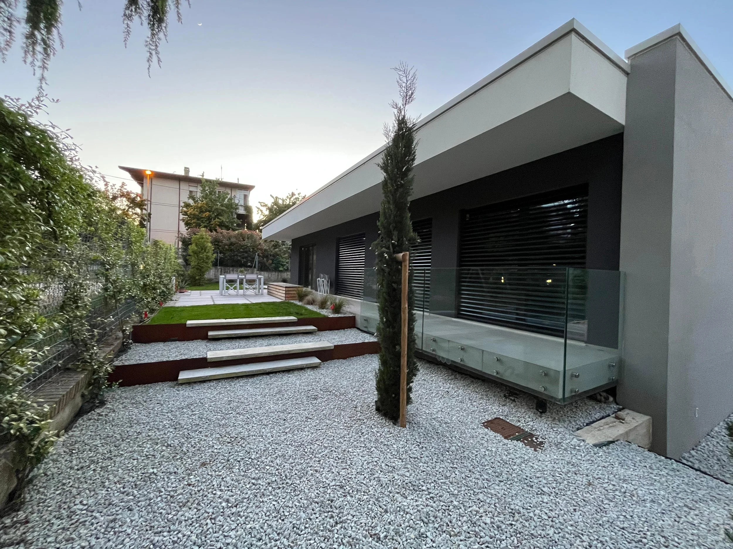 Cortile privato di villa dal design contemporaneo con pavimentazione in ghiaia bianca e alberature verticali. Progetto di DAT Architetti.