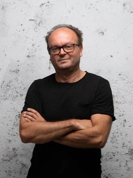 Uomo con capelli grigi, occhiali e maglietta nera, che si appoggia a una parete bianca con texture.