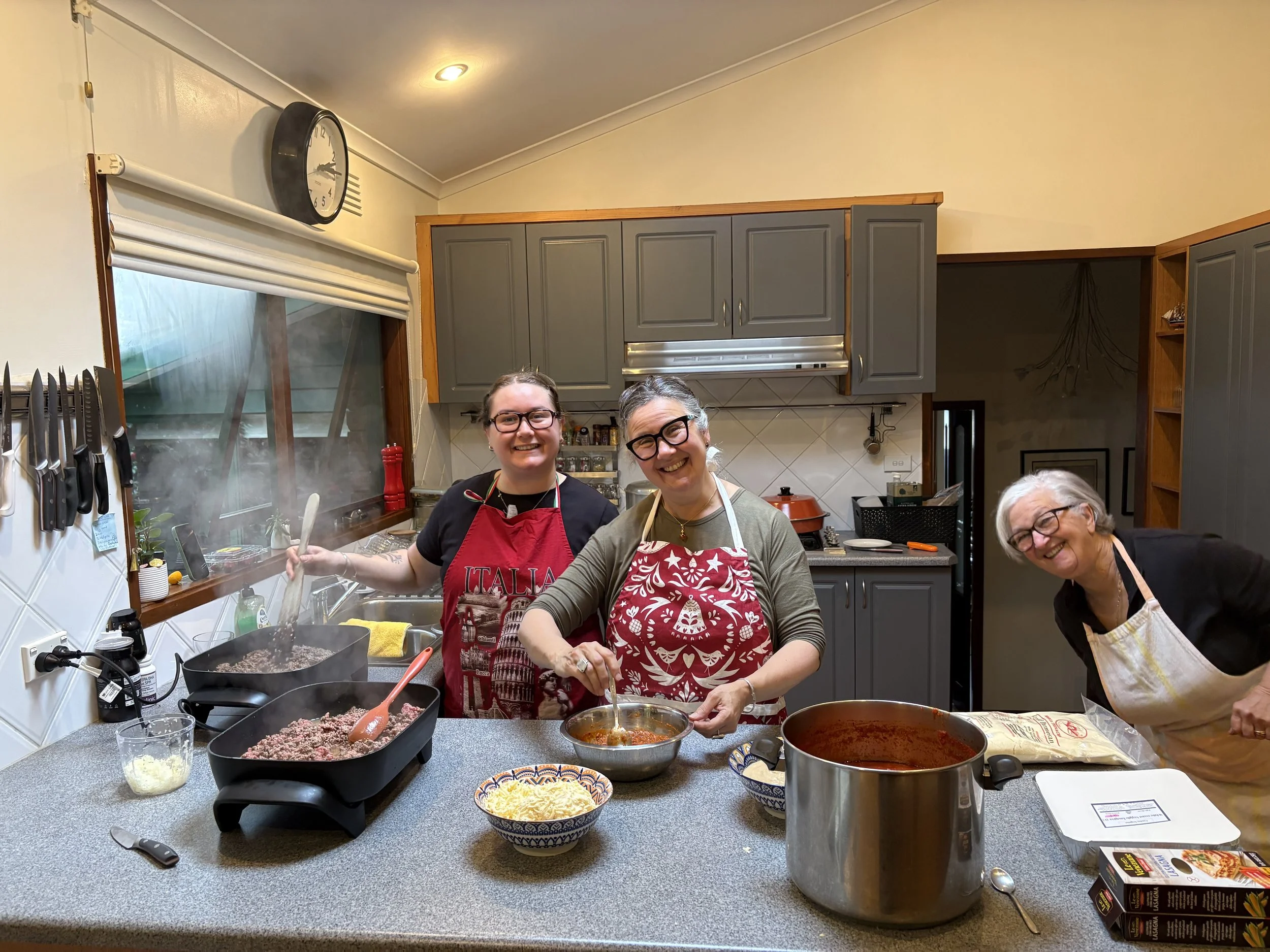 Three women smiling and cooking together in a kitchen, wearing aprons, with various cooking utensils, ingredients, and pots on the counter.