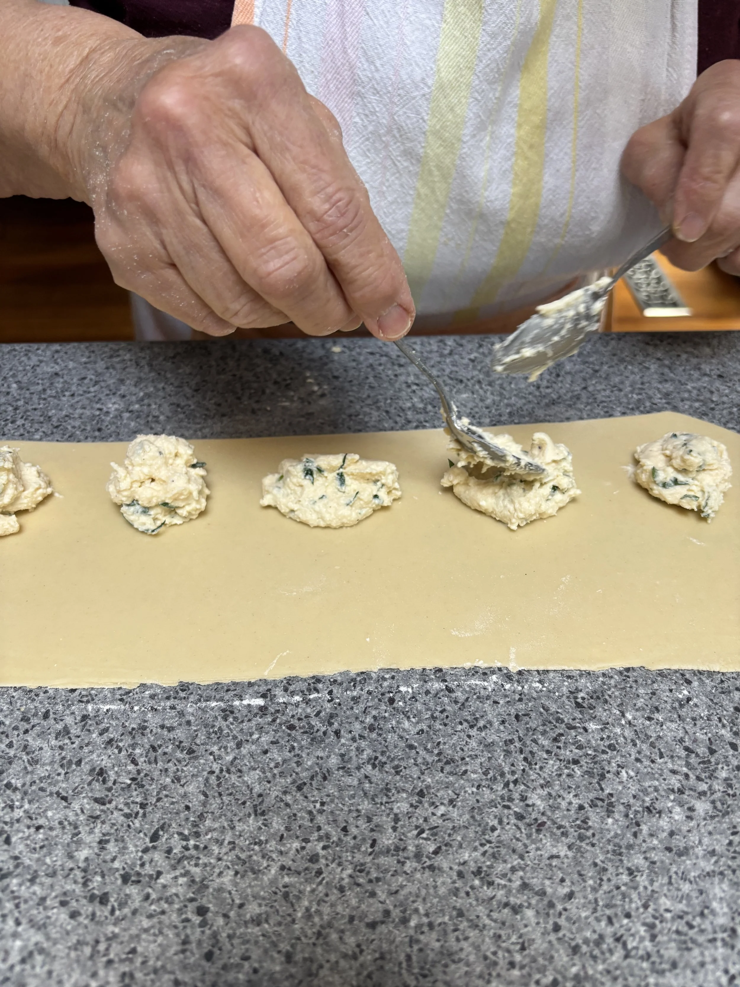 An elderly person uses two spoons to scoop cookie dough onto a piece of parchment paper on a kitchen counter. The person wears an apron, and the dough contains visible chocolate chips and green pieces.