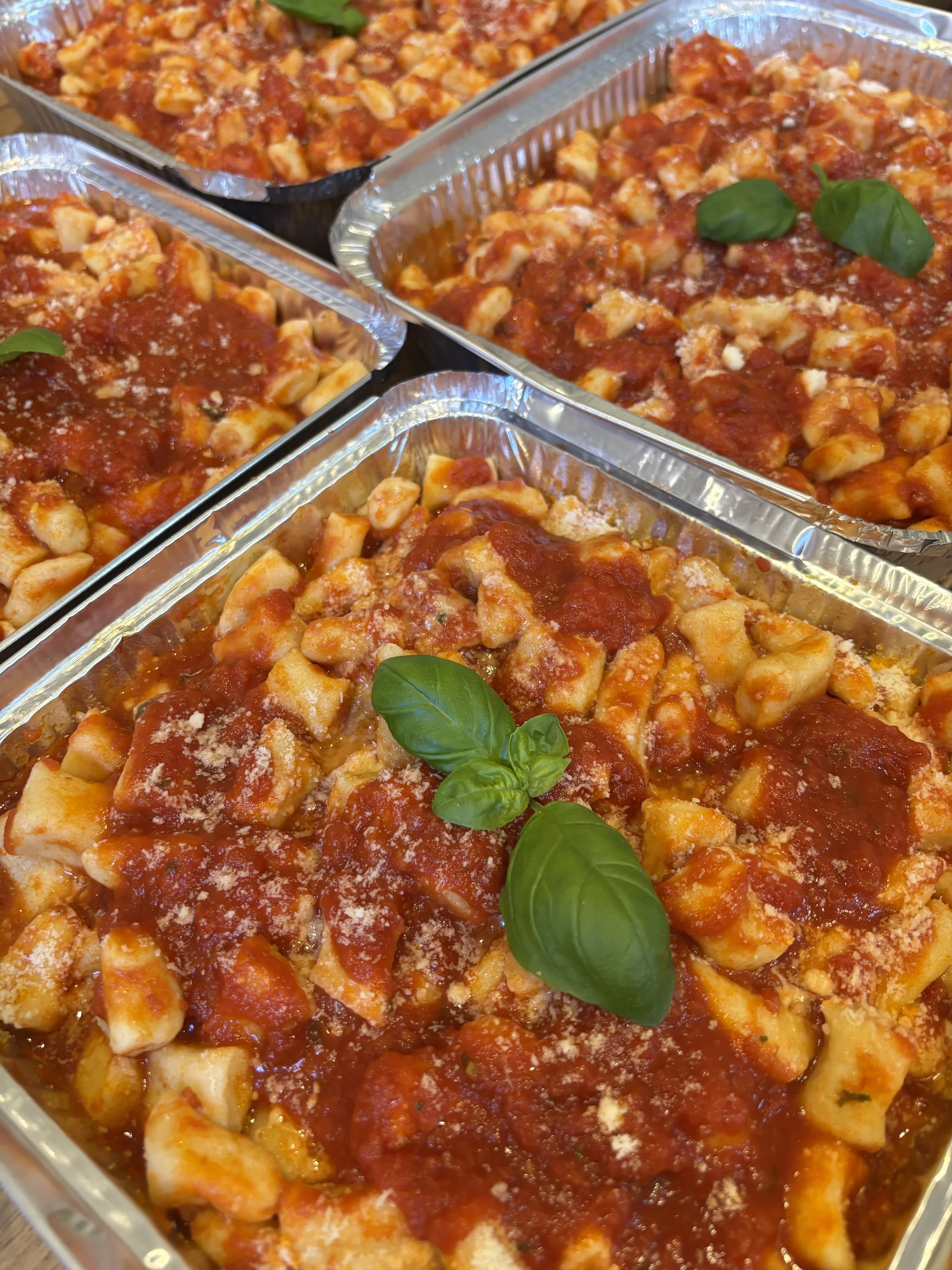 Four aluminum trays filled with uncooked gnocchi pasta topped with tomato sauce, garnished with fresh basil leaves and sprinkled with grated cheese.