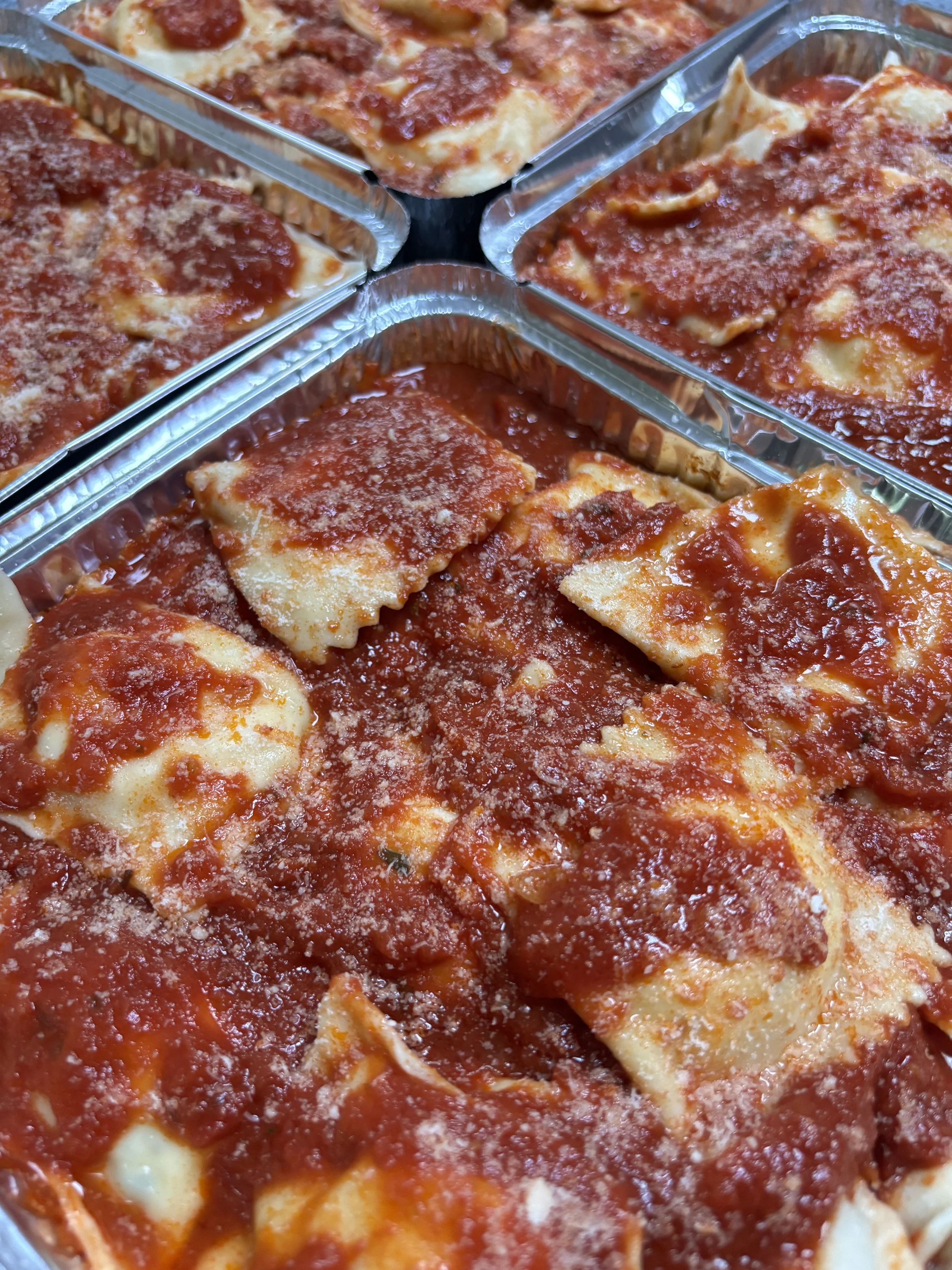 Four aluminum trays of uncooked oven-ready baked cheese pizza with tomato sauce and shredded cheese.