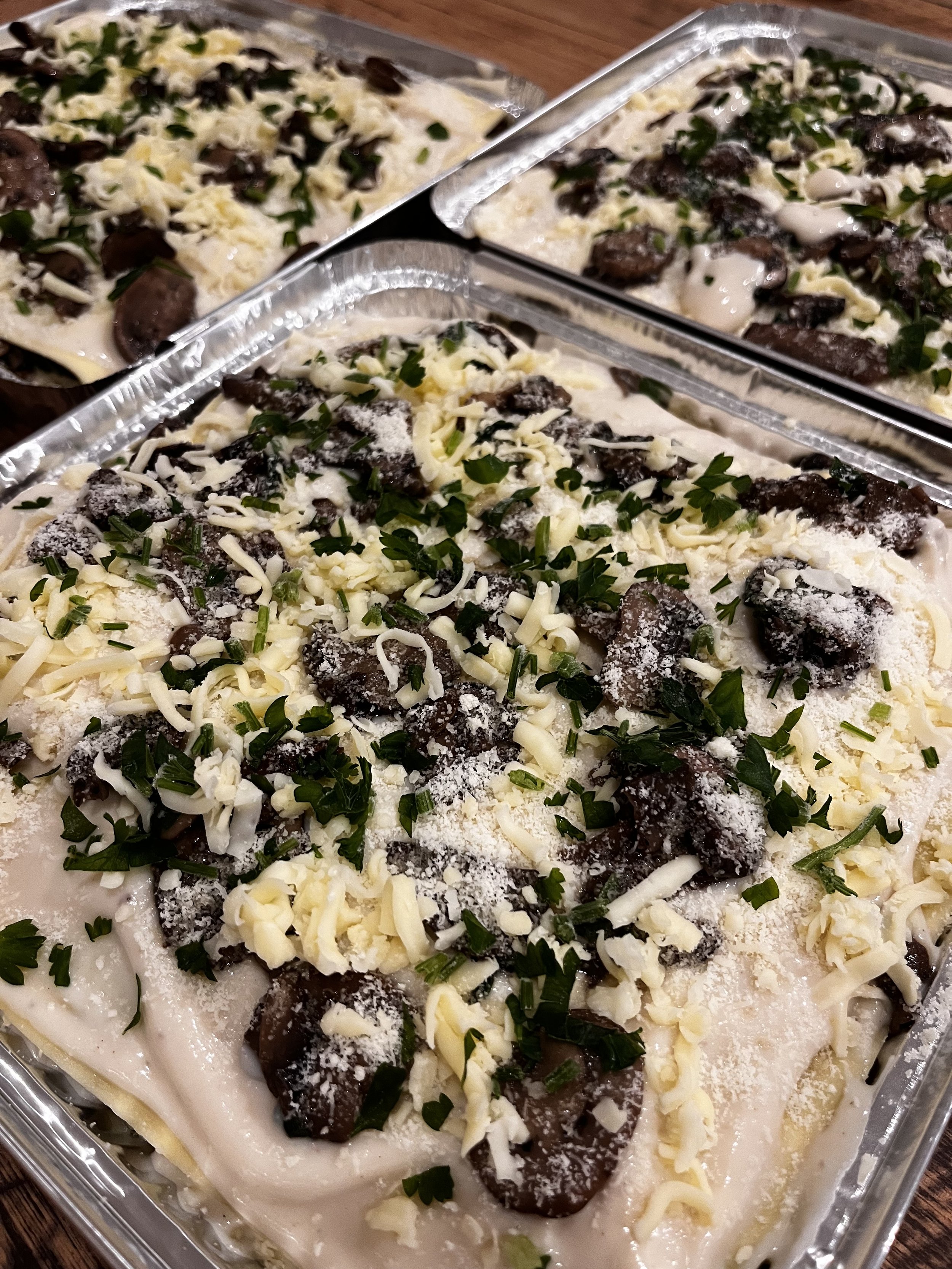 Unbaked lasagna with layers of creamy sauce, mushrooms, shredded cheese, chopped herbs, and spinach in a metal baking dish.