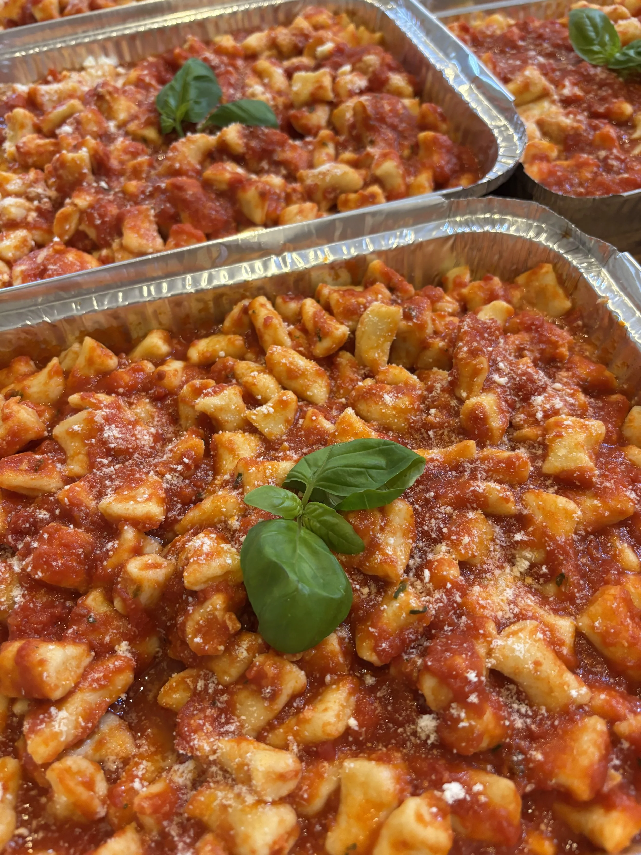 Three foil containers filled with cooked chicken pasta in tomato sauce, garnished with basil leaves and grated cheese.