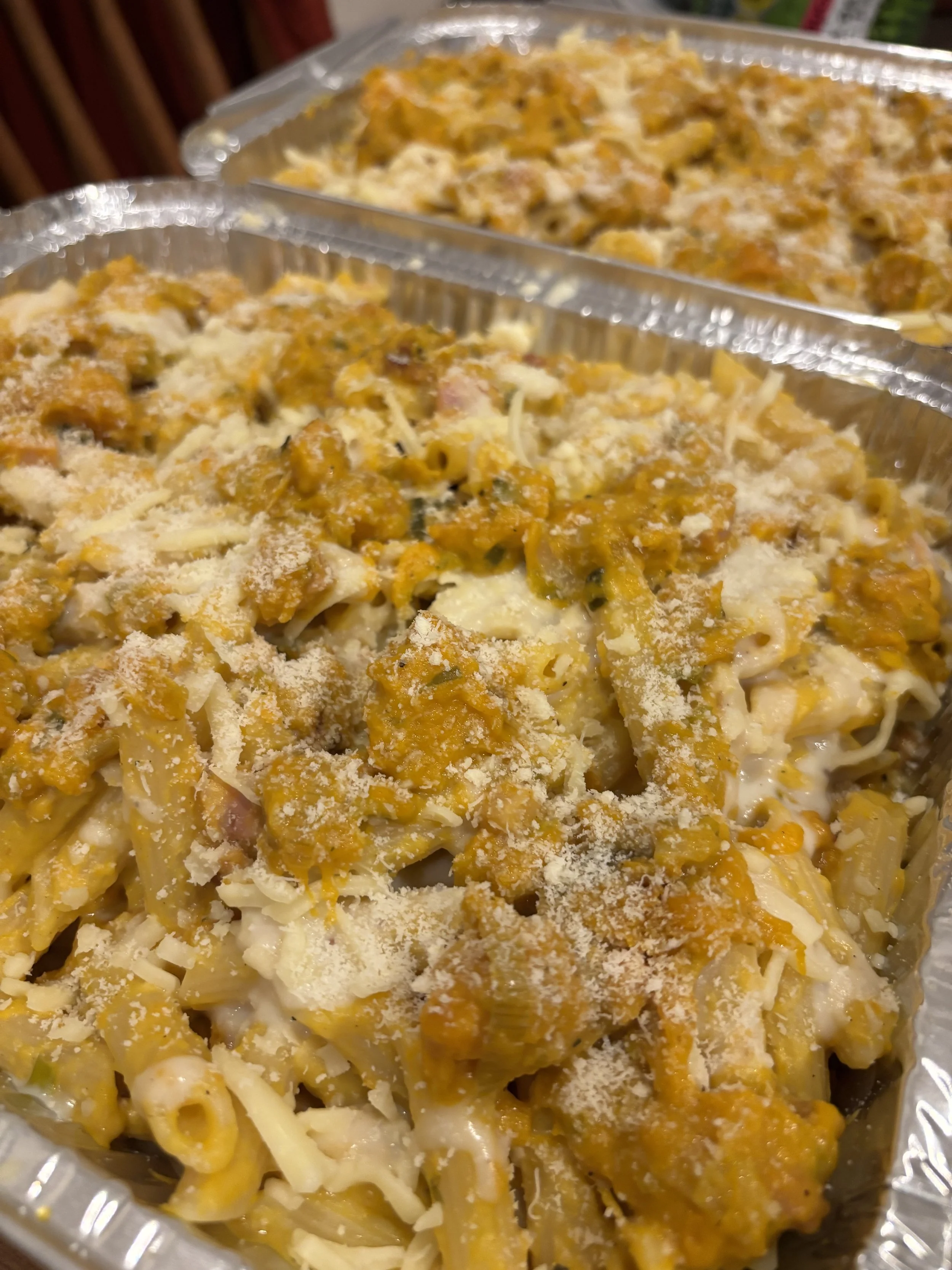 Two aluminum trays filled with baked pasta casserole topped with cheese and grated cheese.
