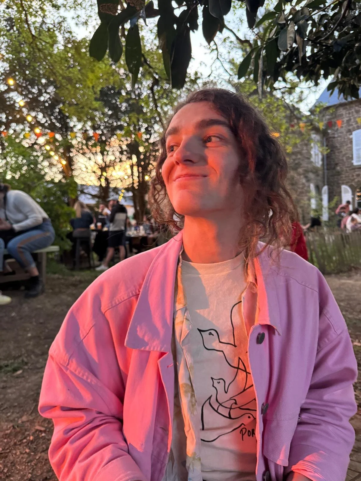 A person with curly hair wearing a pink jacket and graphic t-shirt, smiling and looking to the left, with an outdoor gathering and string lights in the background during sunset.