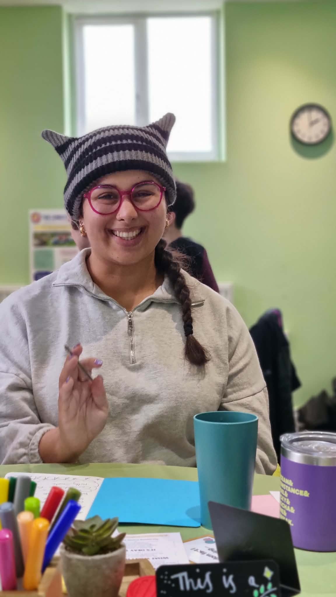 A young woman wearing a striped knit hat with cat-like ears, pink glasses, and a light-colored sweatshirt smiling at the camera. She is sitting at a table with colorful markers, a small plant, a blue cup, and various papers.