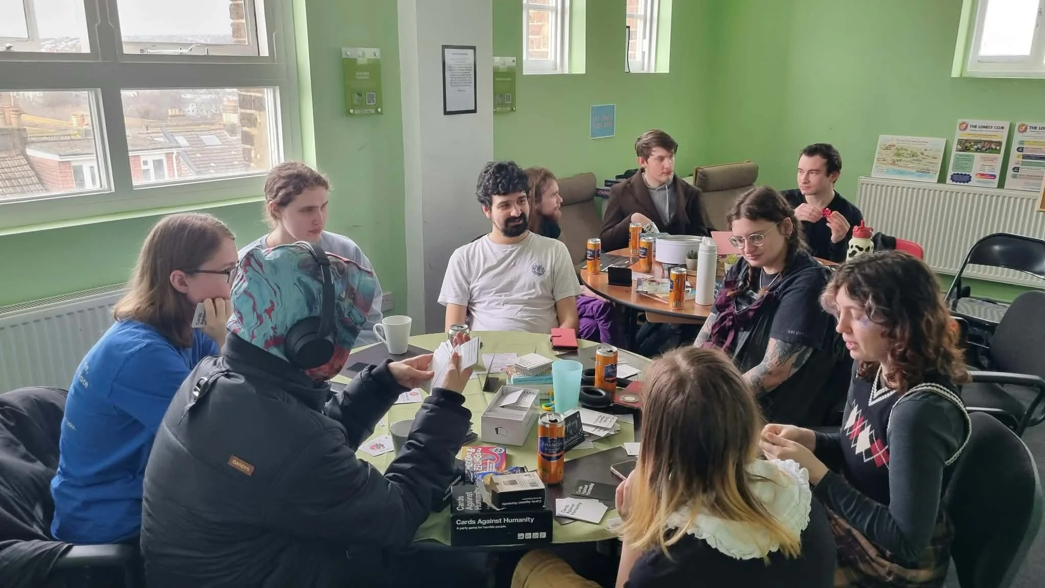 A group of people sitting around a table playing a card game in a room with green walls and large windows.