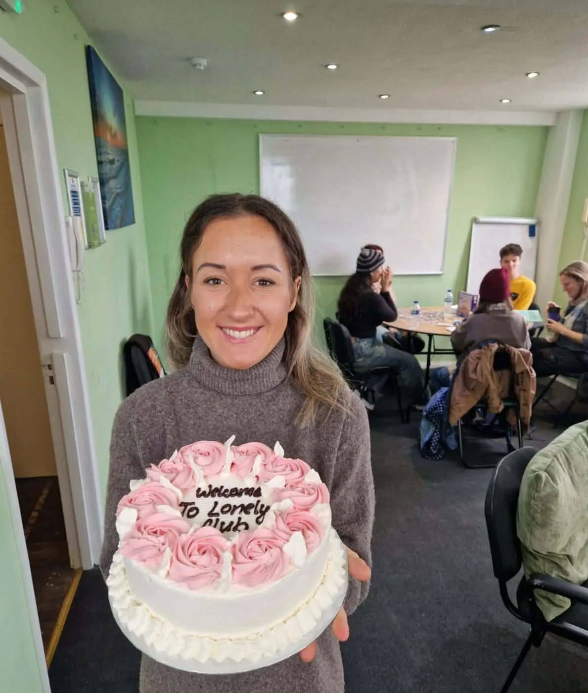 A woman holding a birthday cake with pink roses and the message 'Wishing You Lots of Love' written on it. In the background, several people are sitting at tables in a green-walled room.