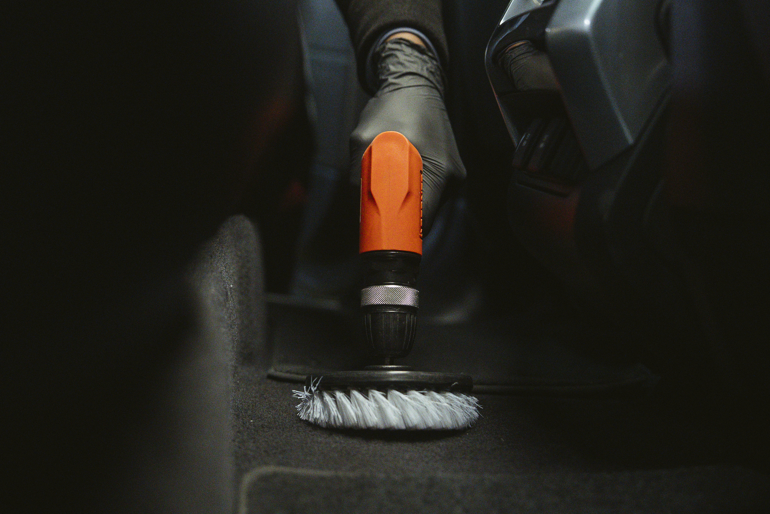 A person using a cordless power drill with an orange and black body to clean the floor with a round brush attachment in a vehicle interior.