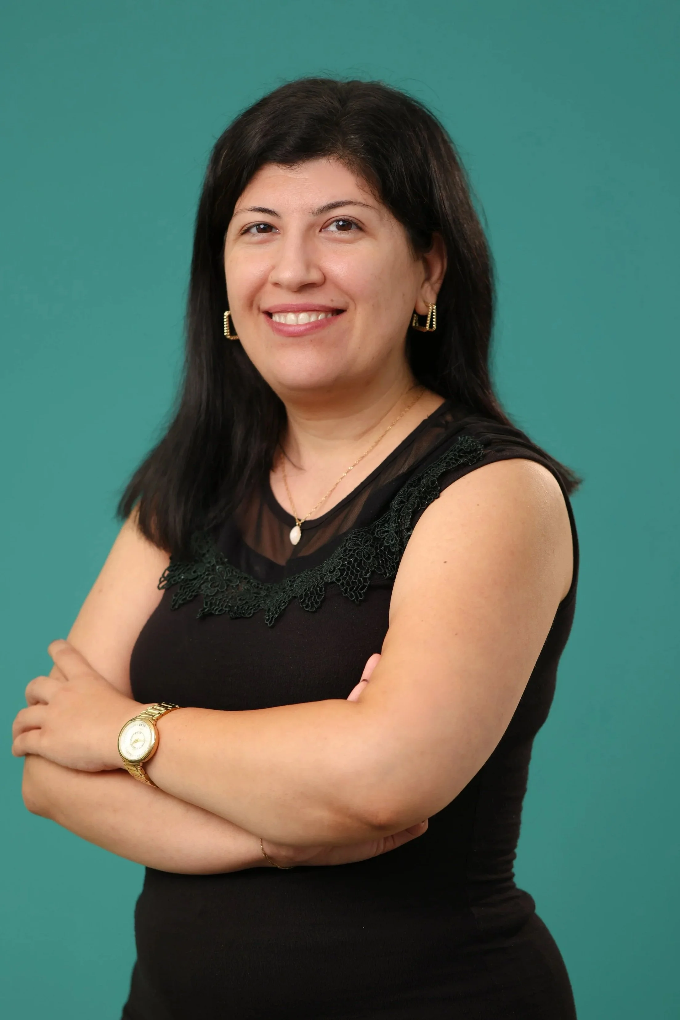 A woman with dark hair smiling, wearing a black sleeveless dress with lace detail, gold earrings, a necklace with a white pendant, and a gold watch, standing against a teal background.