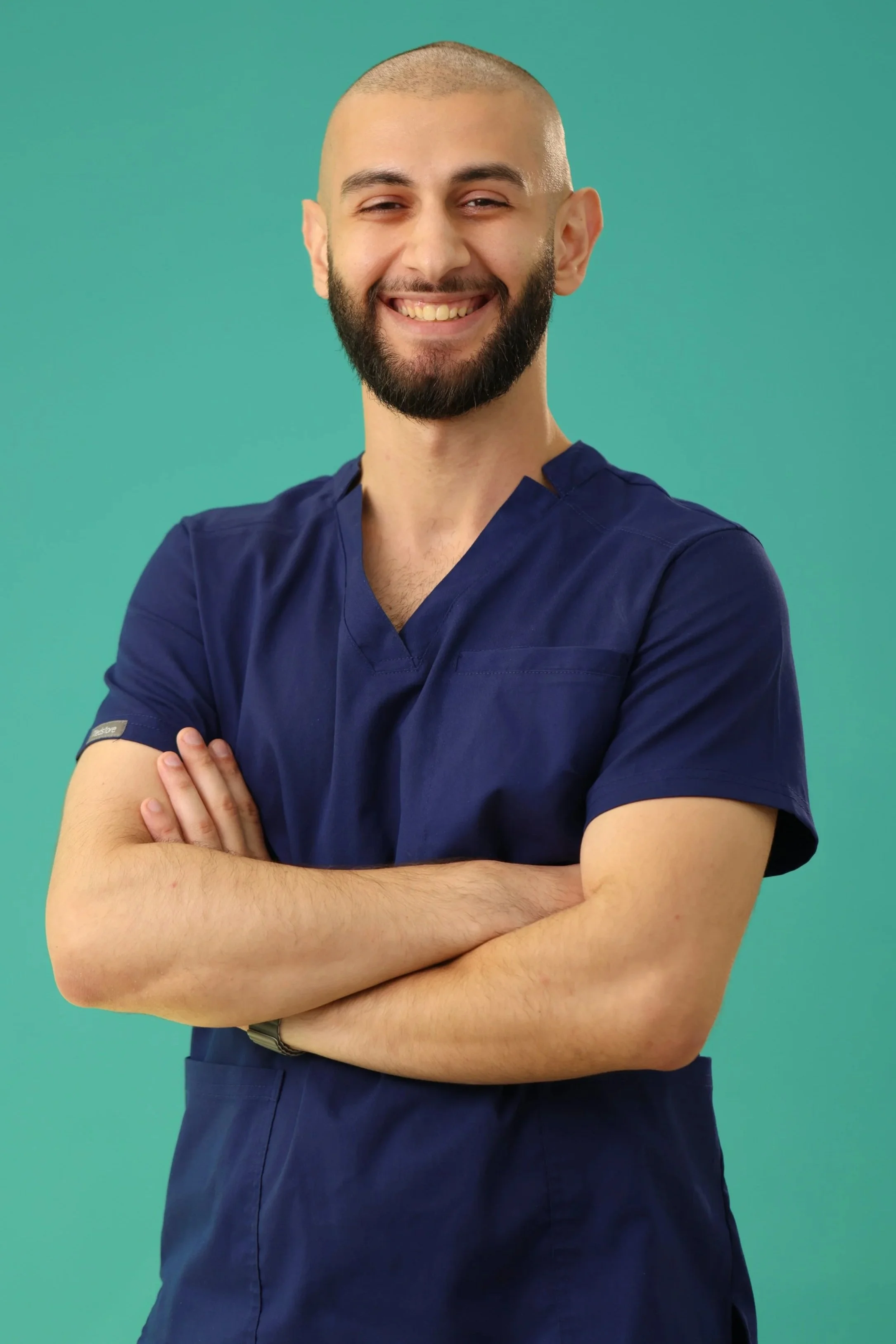 Smiling male healthcare professional with crossed arms wearing navy scrubs against a green background.