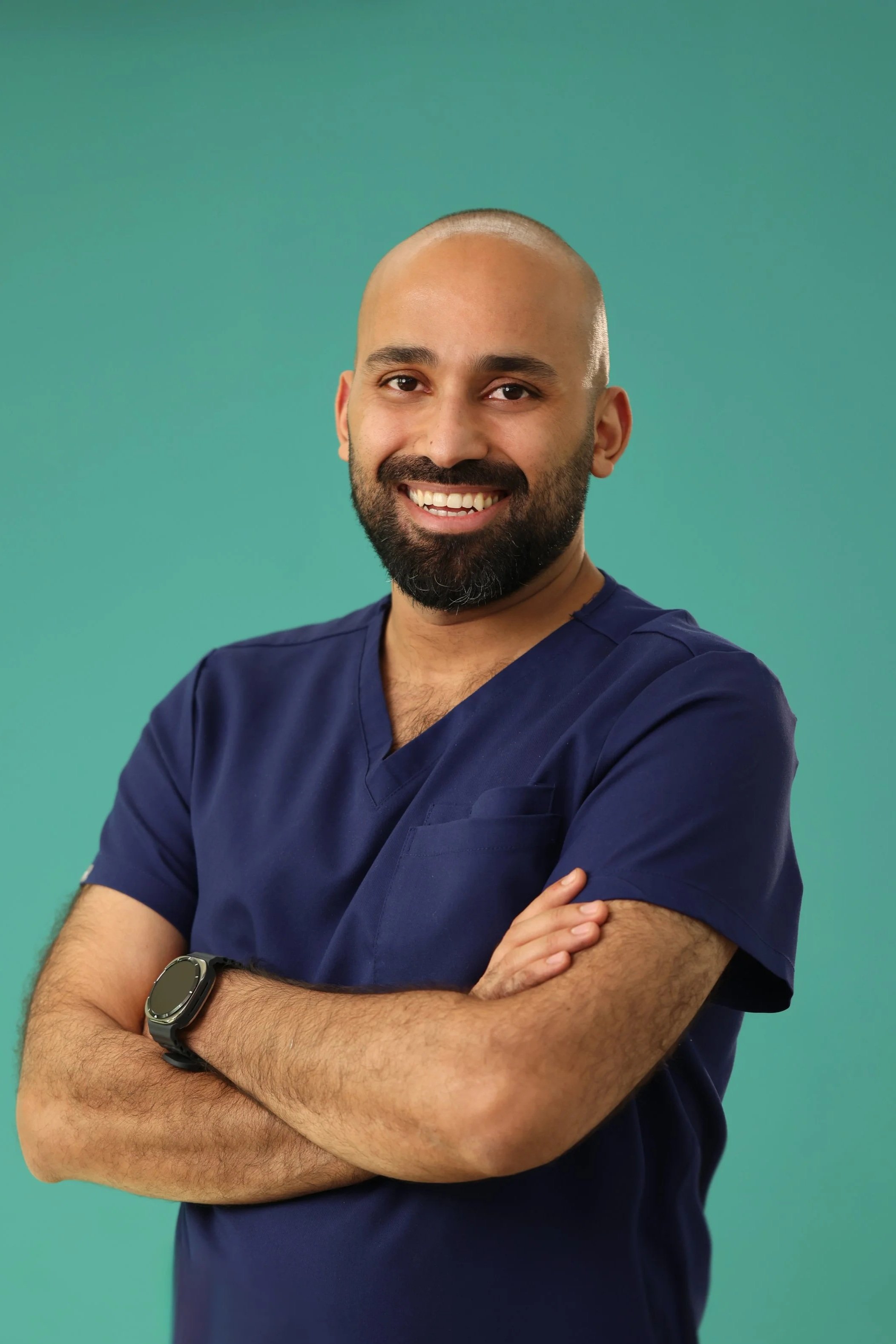 Portrait of a smiling man with a shaved head and beard, wearing navy medical scrubs and a smartwatch, standing with arms crossed against a green background.