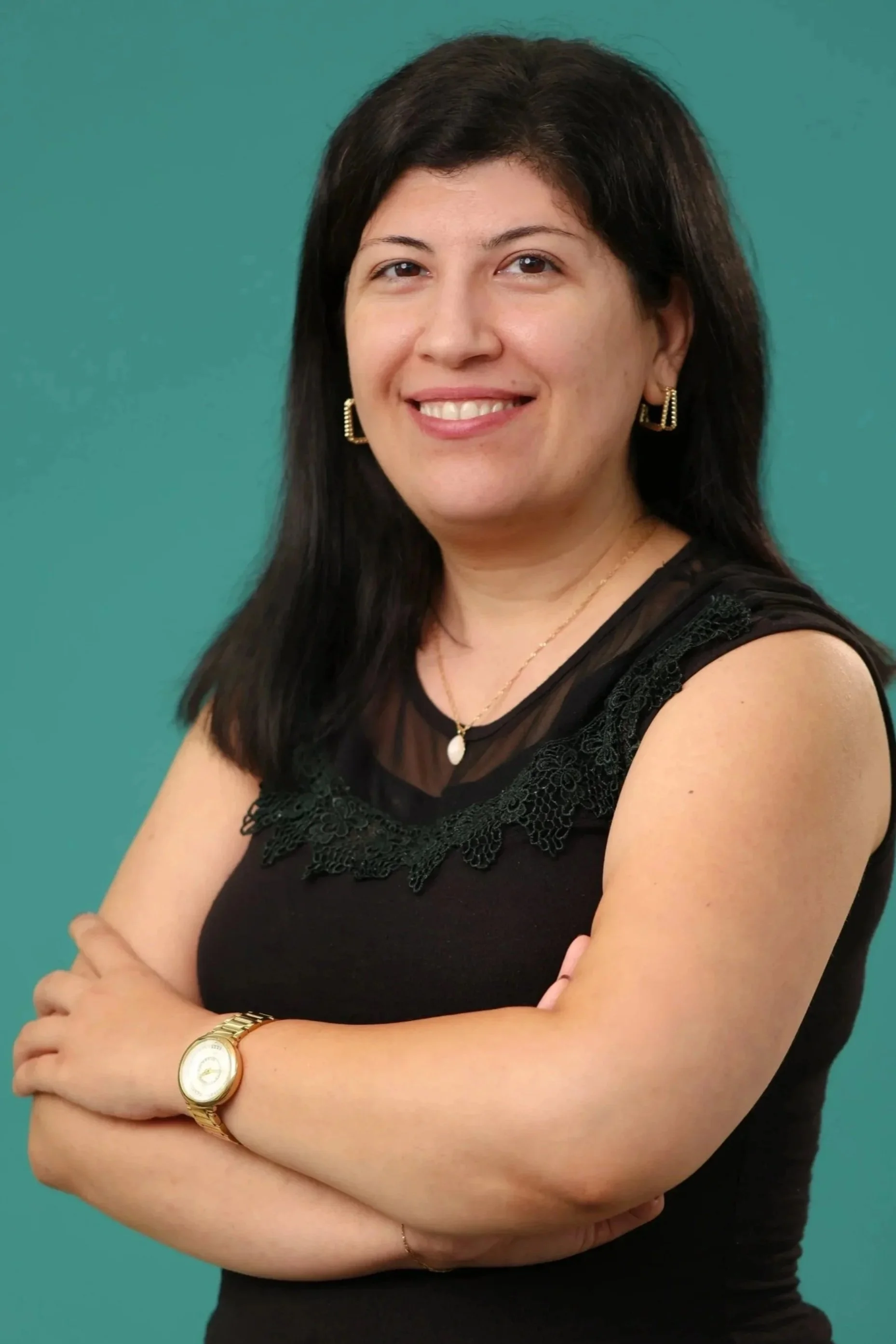 A woman with black hair smiling at the camera, wearing a black top with lace detail, gold earrings, a necklace, a gold watch, and a gold bracelet, standing with her arms crossed against a green background.