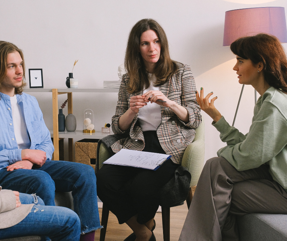 Three women and a man having a conversation in a living room, with a shelf and a pink lamp in the background.