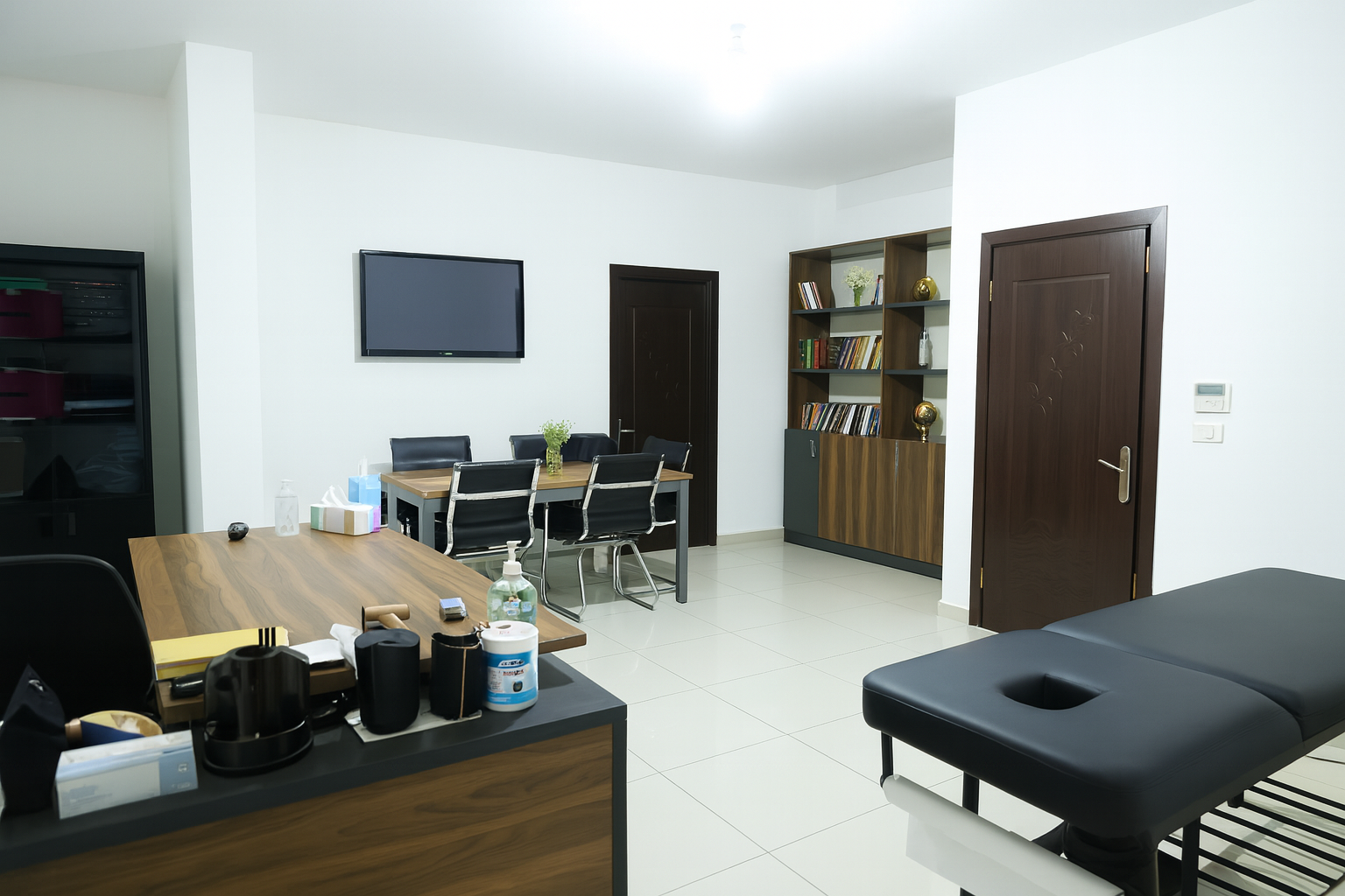 Medical office or examination room with a black massage table, a wooden desk with various items, a dining table with six chairs, a wall-mounted TV, a bookshelf with books and decorative items, and a closed wooden door.