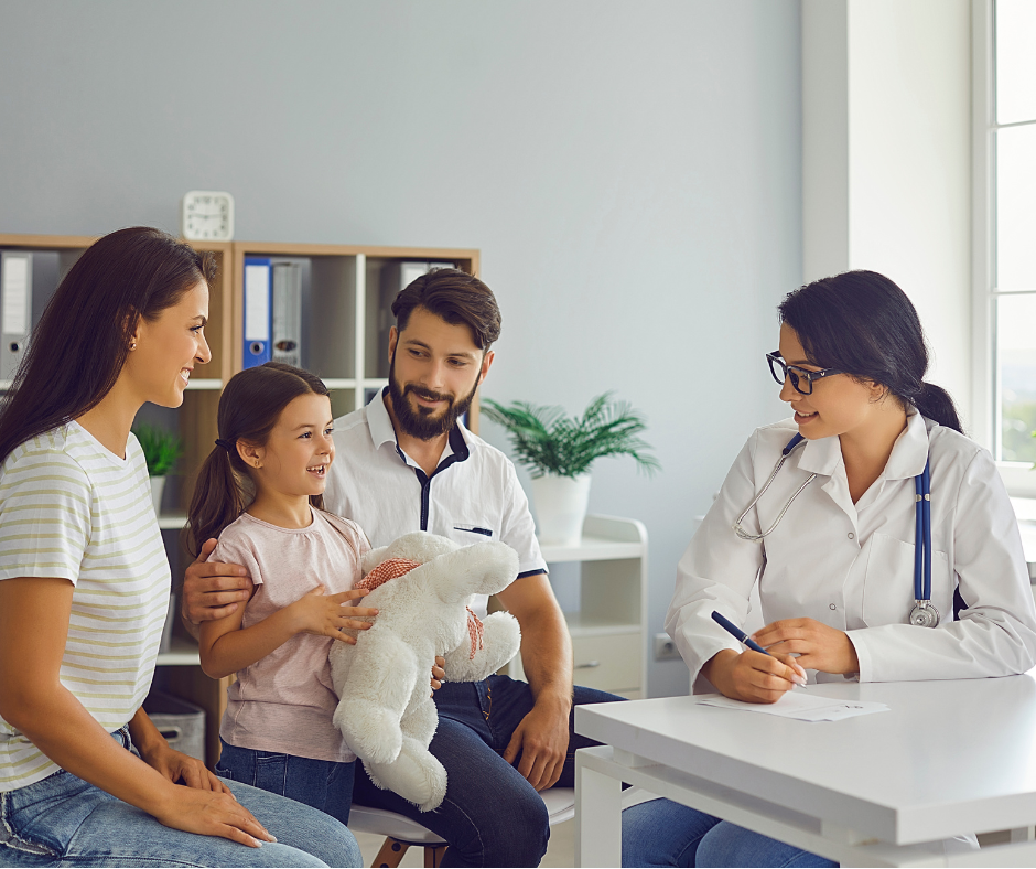 A young girl holding a stuffed animal talking to a female doctor at a medical consultation, with her parents sitting beside her in a bright doctor's office.