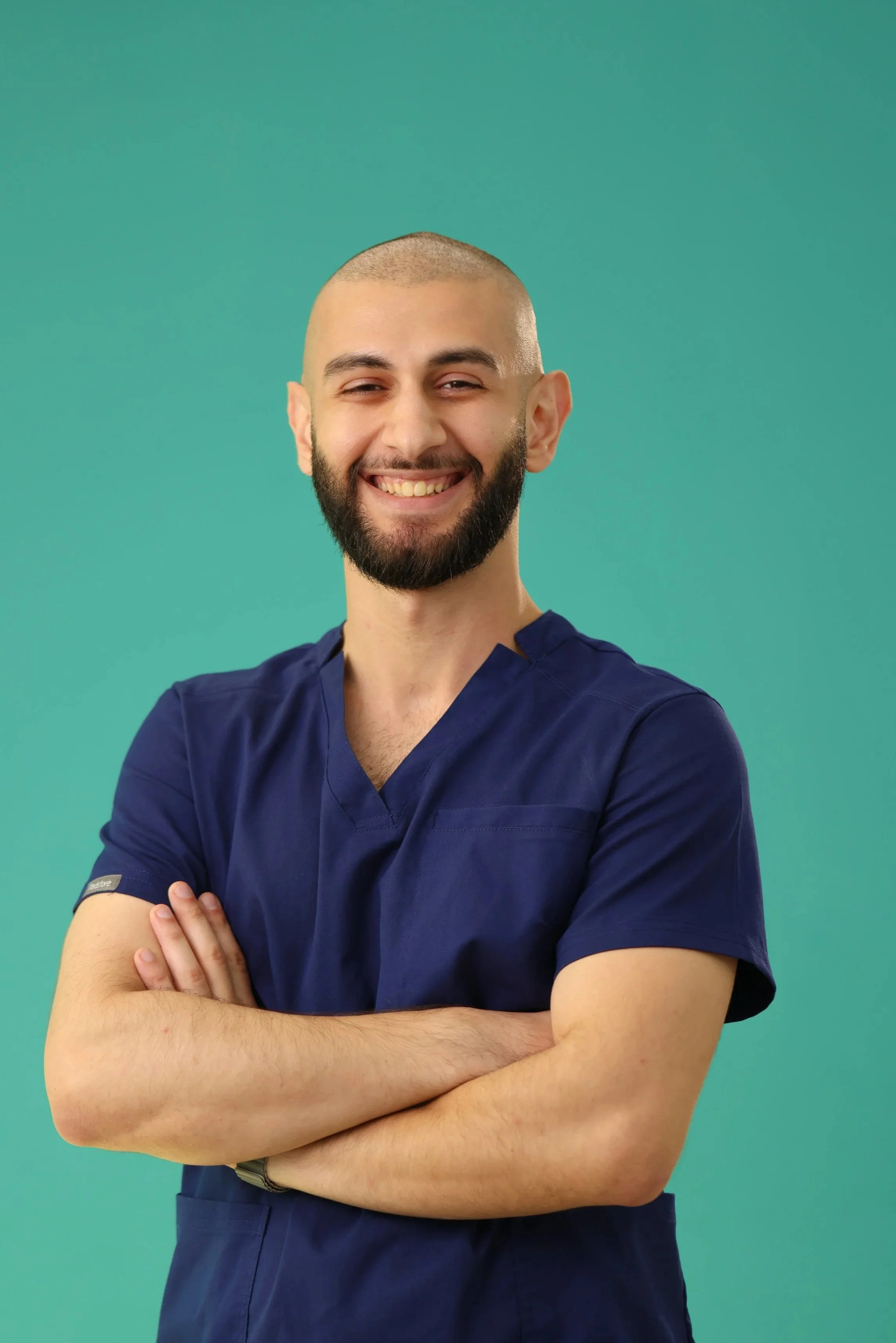Smiling man in navy scrubs with crossed arms against a teal background.