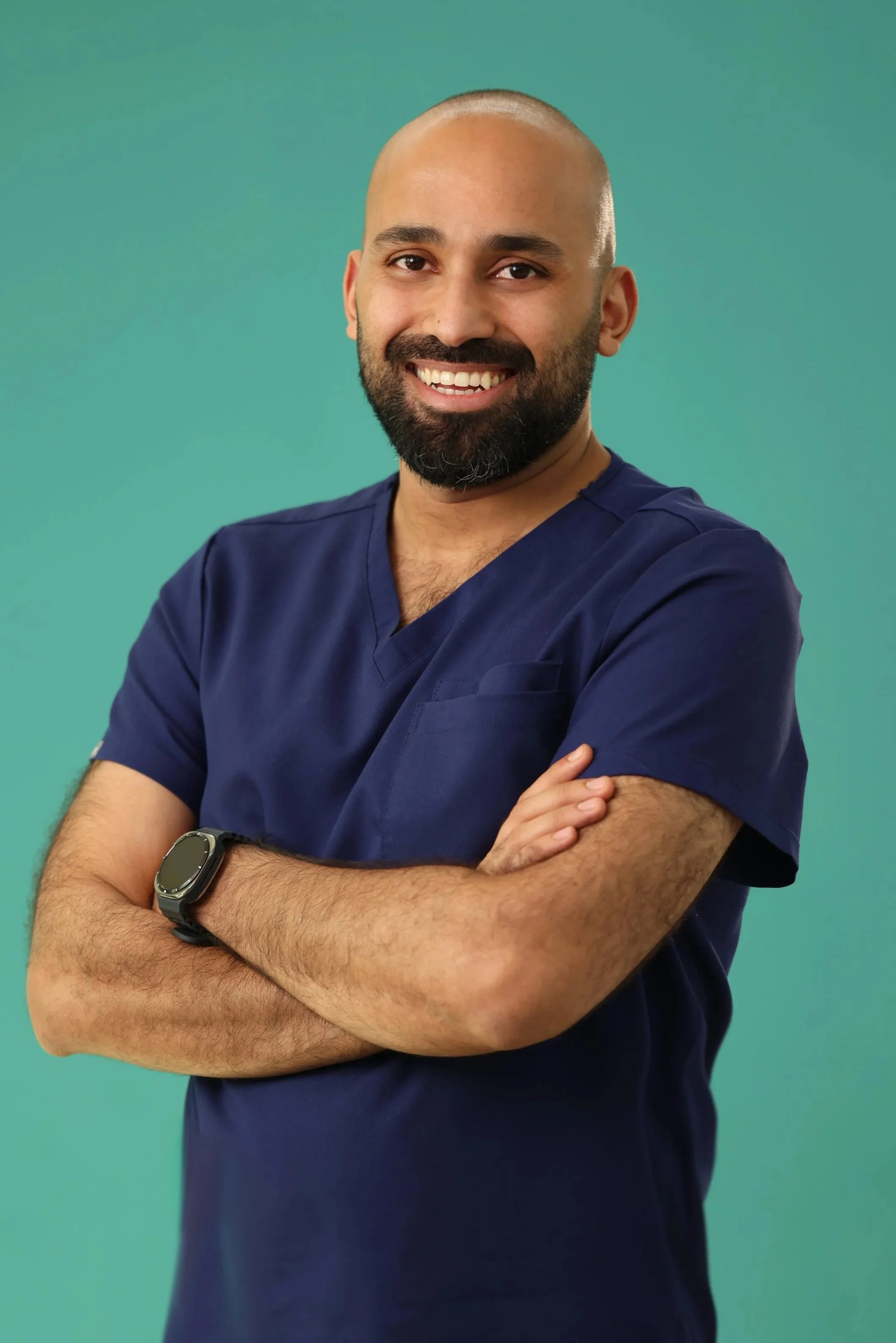 A smiling man wearing navy blue medical scrubs with arms crossed, standing against a teal background.