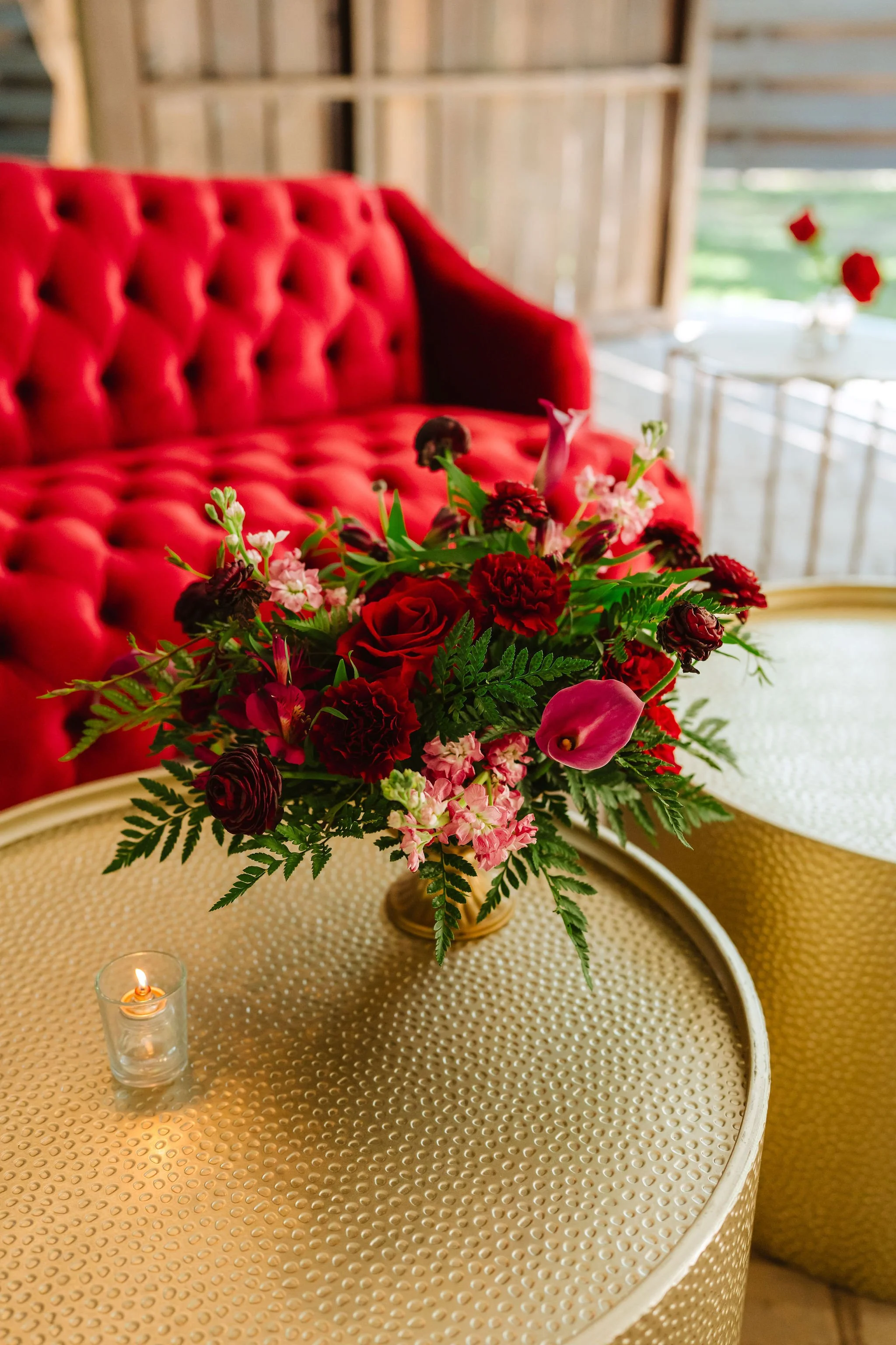 Rich red and burgundy floral arrangement displayed beside a red velvet lounge at an elegant wedding reception.