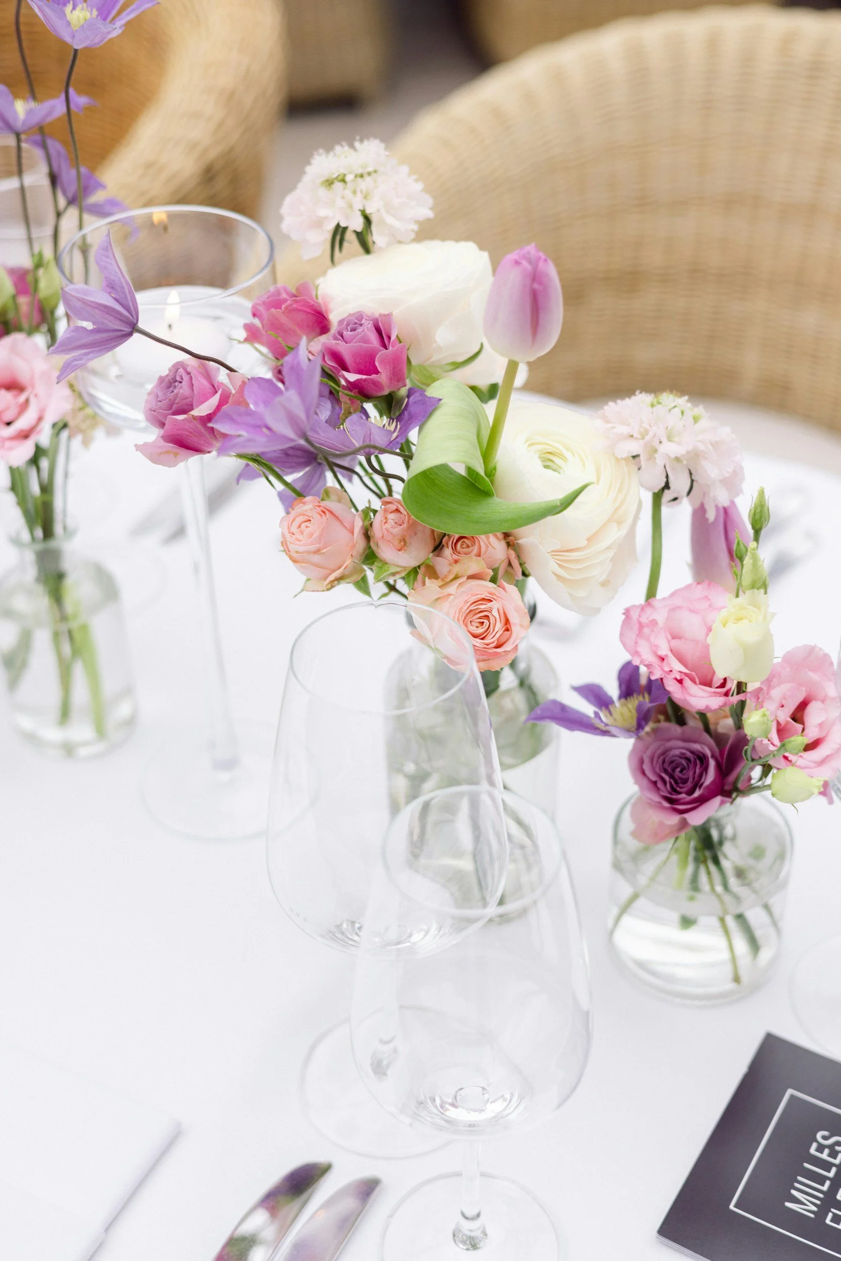 Colorful pink, purple, peach, and white wedding flowers arranged in modern bud vases on a white table.