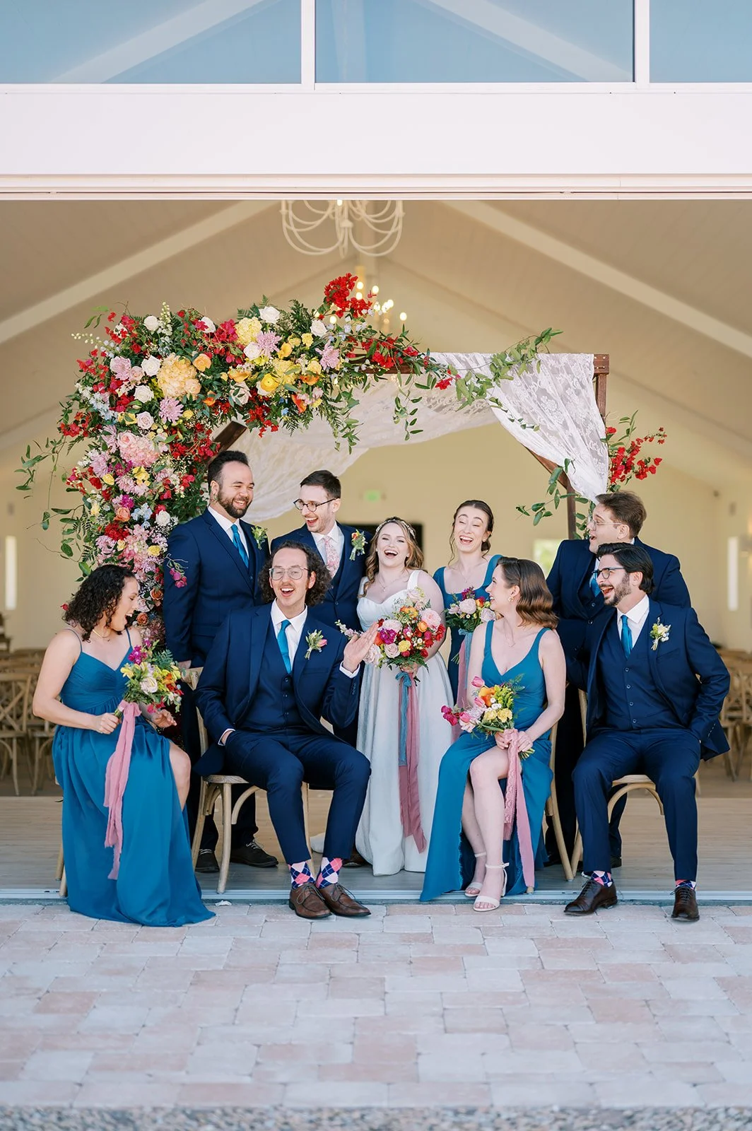 Wedding party in blue and pink attire holding bright colorful bouquets beneath a floral ceremony arch.
