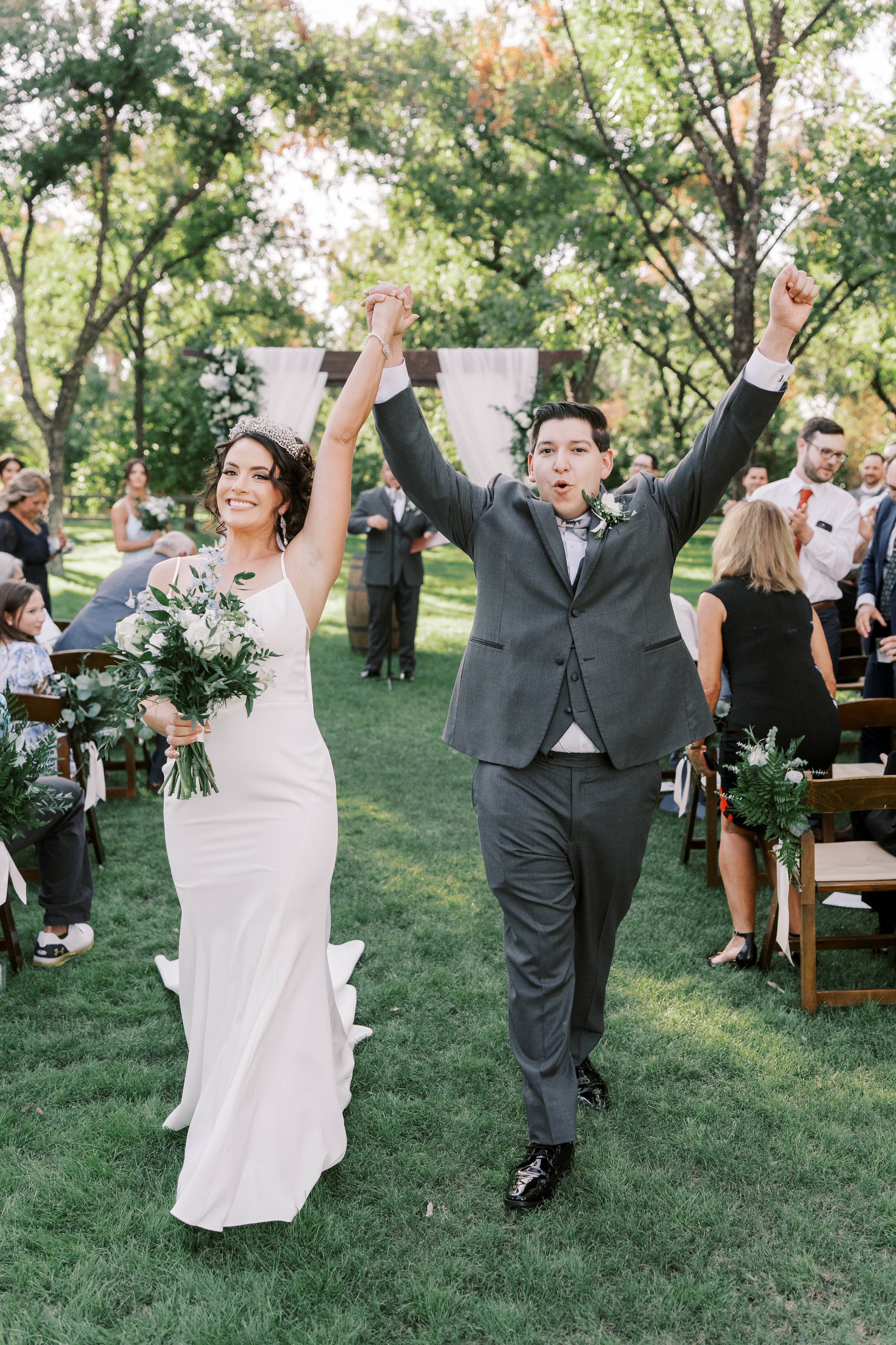 Newlyweds celebrating after their romantic outdoor wedding ceremony surrounded by white flowers and greenery.