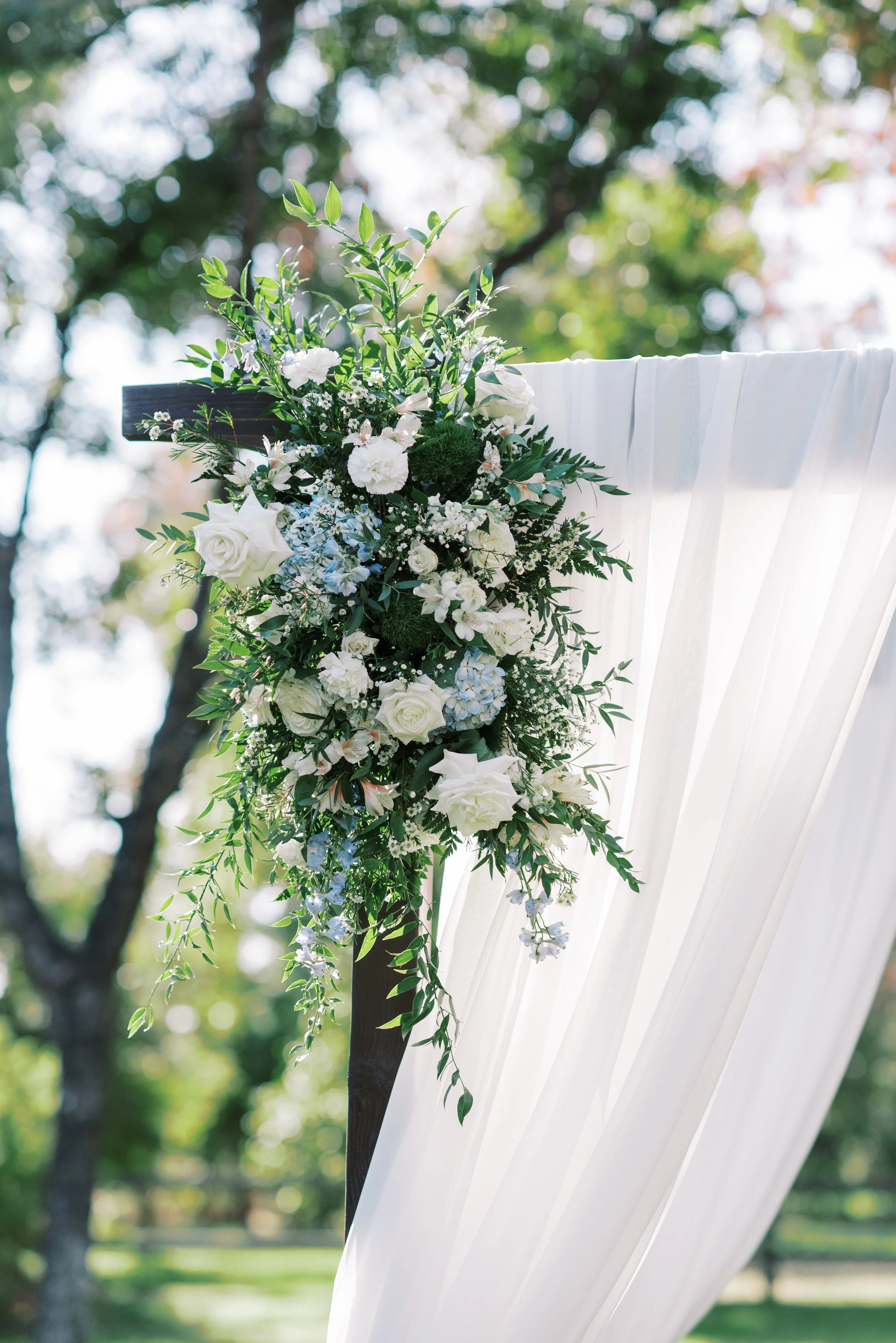 White and blue wedding ceremony floral arrangement with lush greenery on a draped arch at Venue at the Grove.