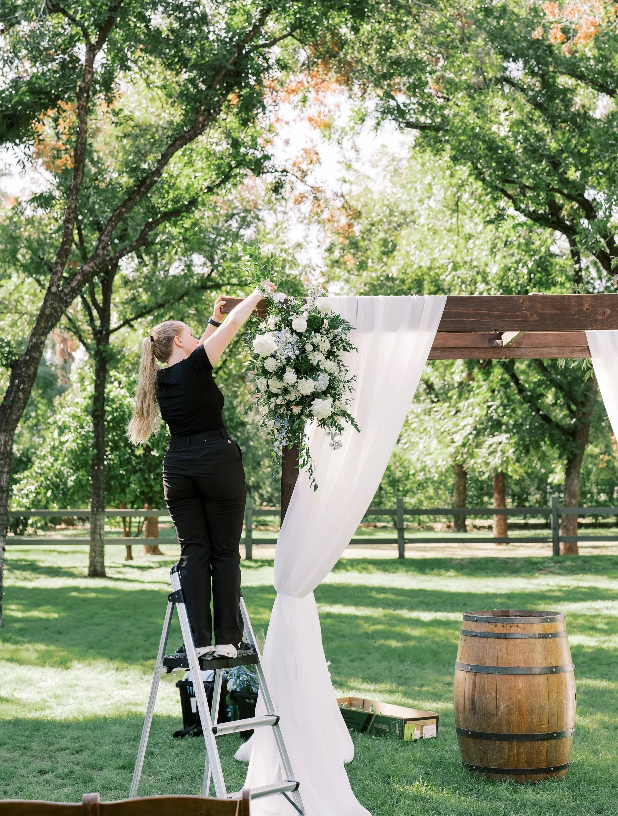 Dani of Bee Wild Designs installing white and green wedding flowers on an outdoor ceremony structure.