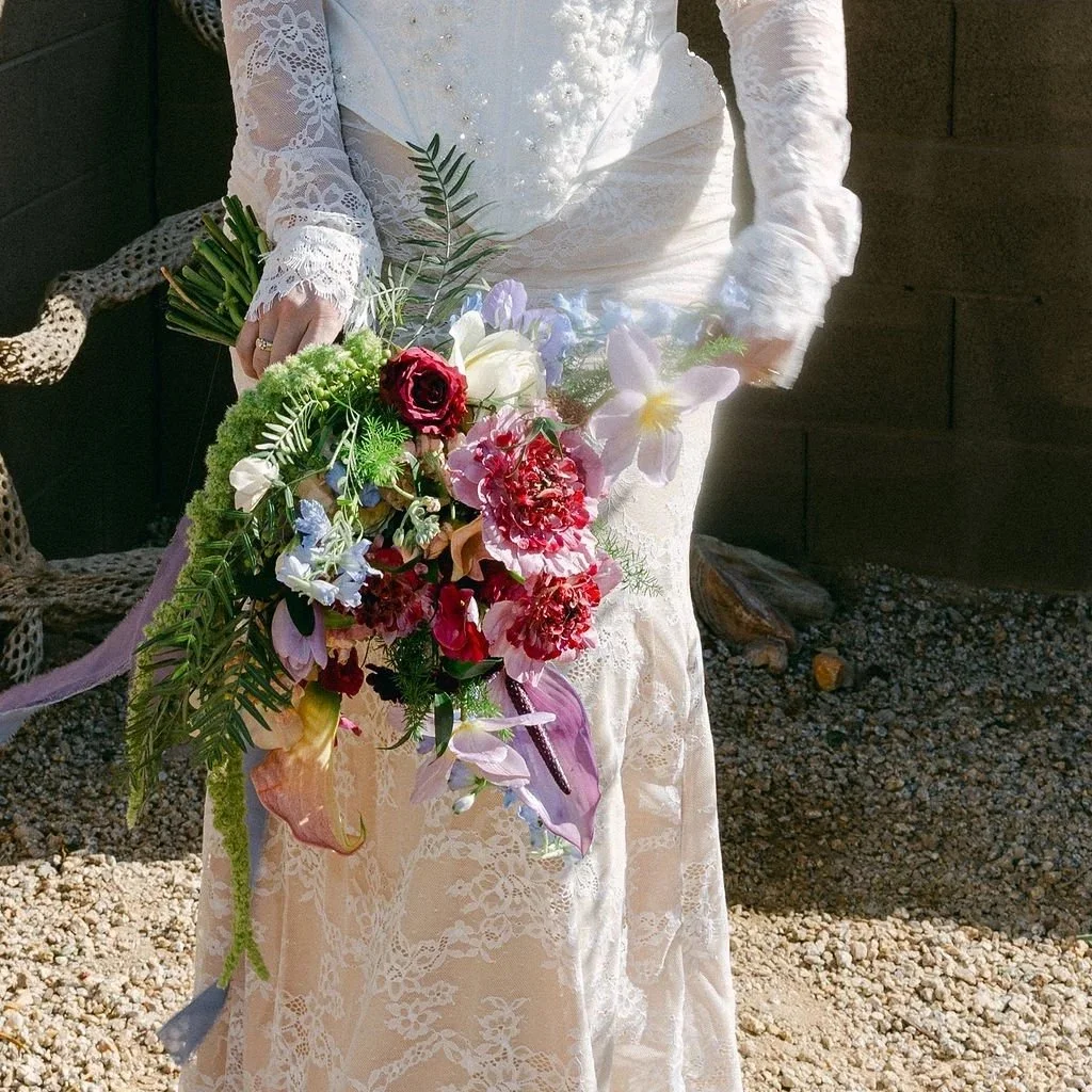 Bride holding a colorful garden-style wedding bouquet with pink, red, blue, white, and textural flowers.