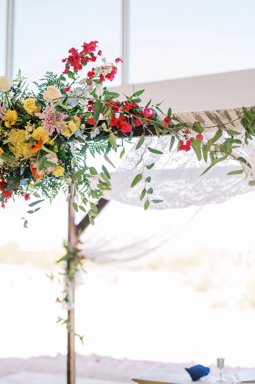 Statement wedding ceremony installation with colorful flowers and greenery cascading across a white draped arch.