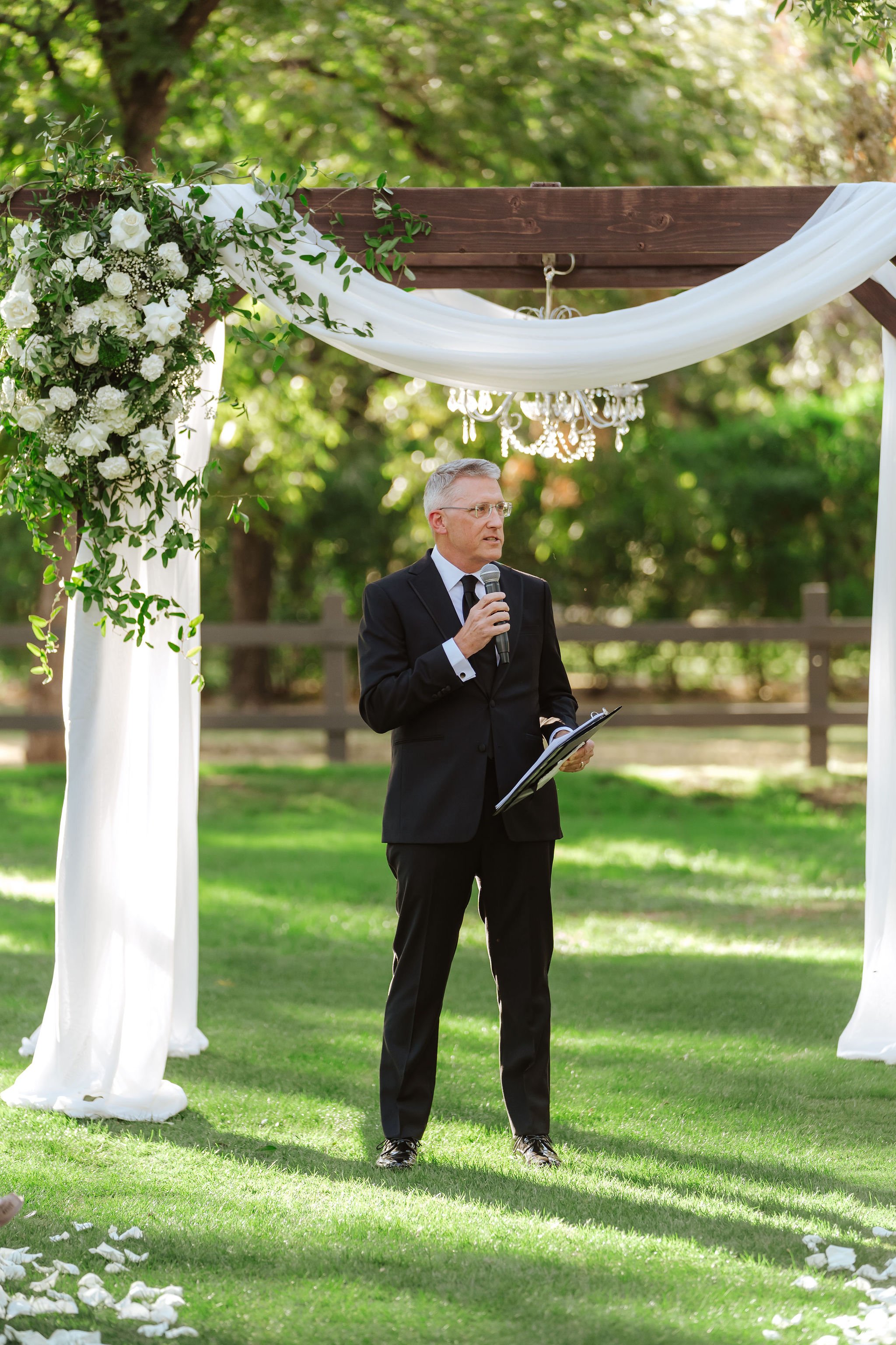 Officiant  standing beside a white-draped outdoor wedding ceremony arch surrounded by trees and greenery.