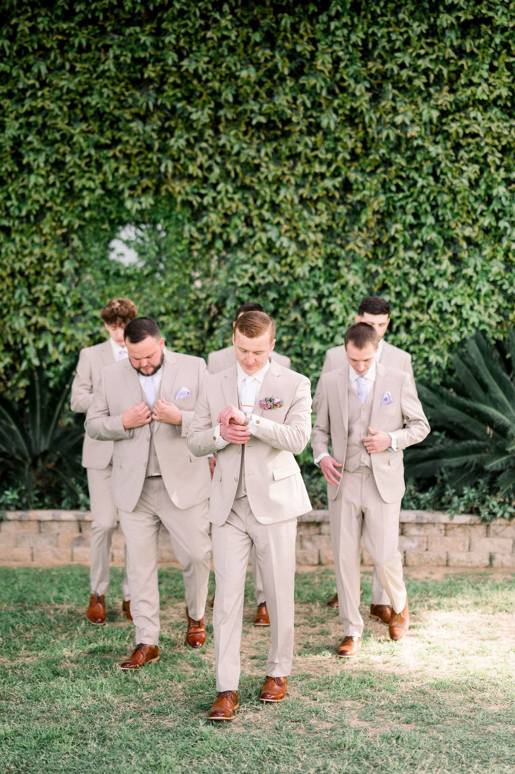 Groomsmen in neutral suits at a rustic Knotty Barn wedding surrounded by lush greenery.