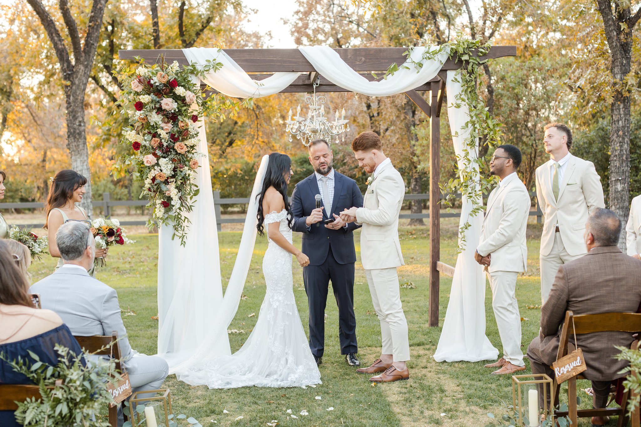 Bride and groom exchanging vows beneath a draped wooden ceremony arch with fall floral installations.