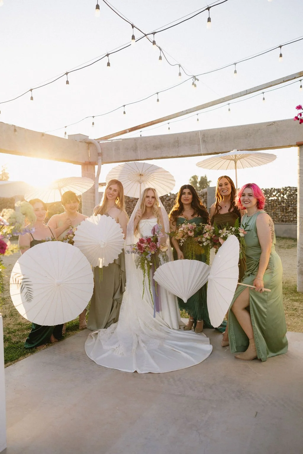 Bride and wedding party gathered outdoors with bouquets beneath a white tent.