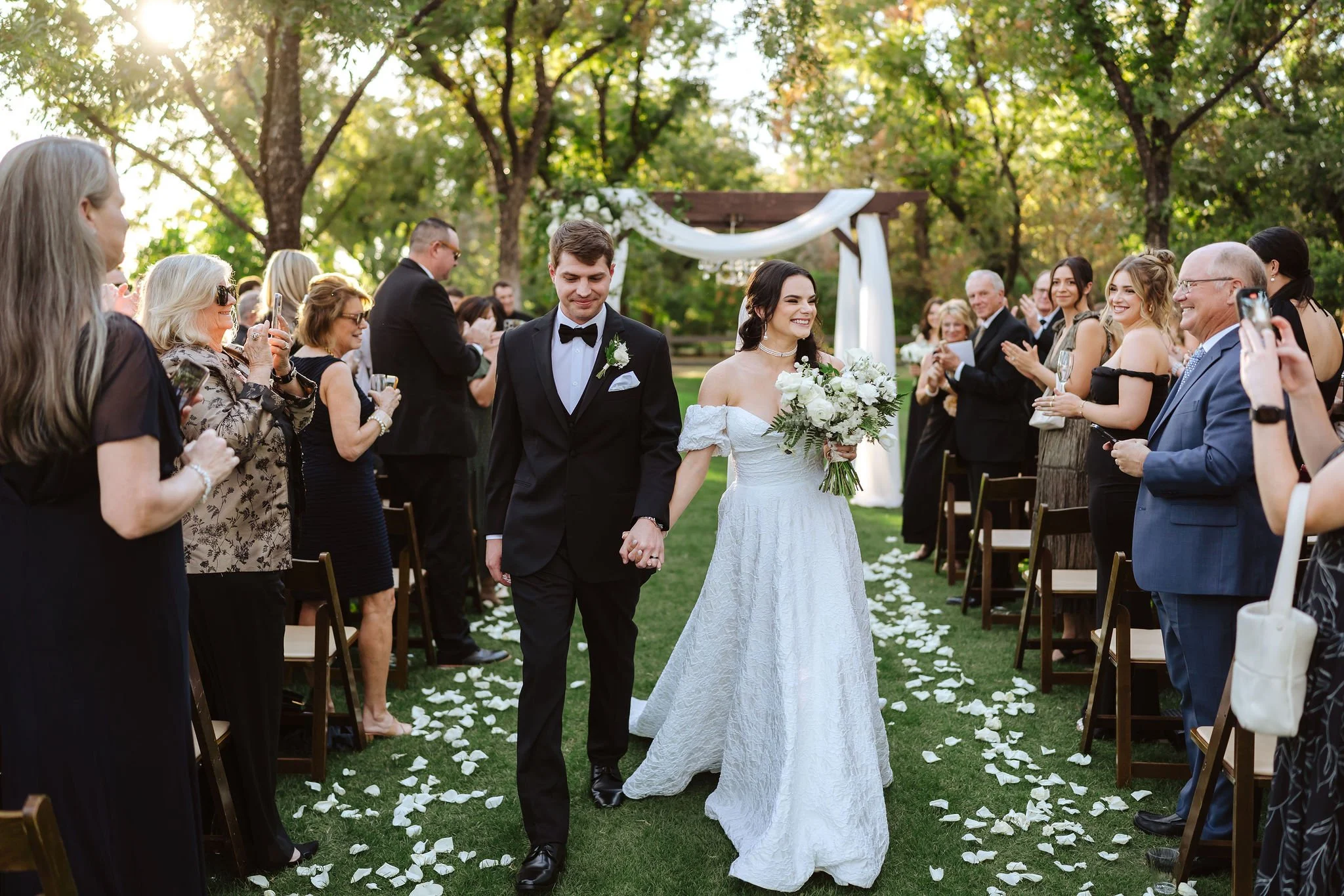 Newlywed couple walking down the aisle after an outdoor wedding ceremony with white floral arrangements and greenery.
