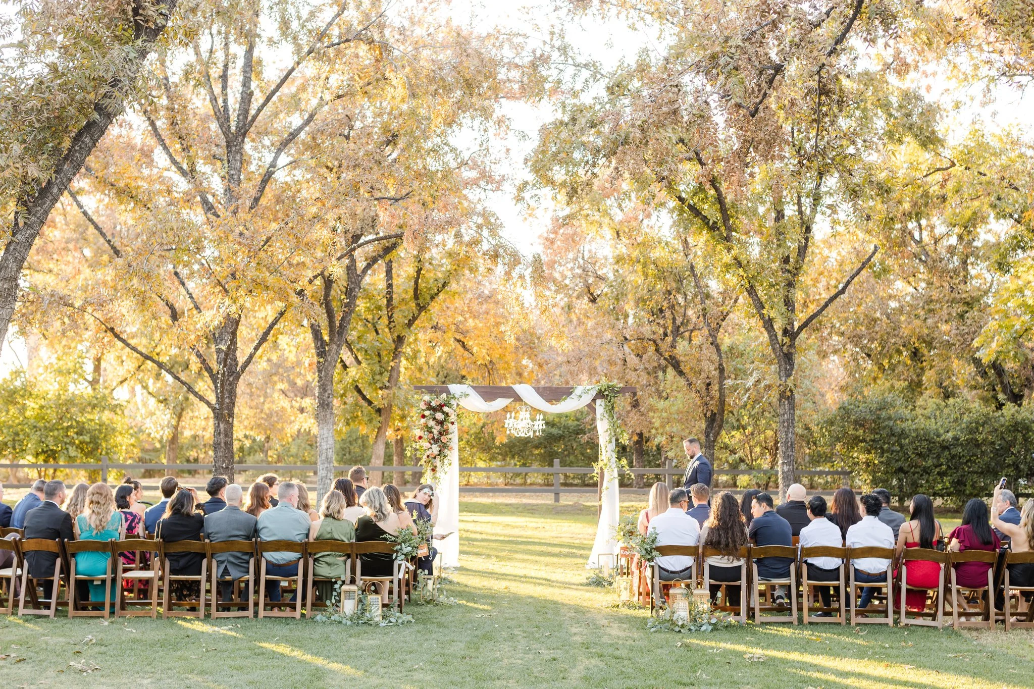 Outdoor fall wedding ceremony beneath mature trees with white draping and garden-inspired floral details.
