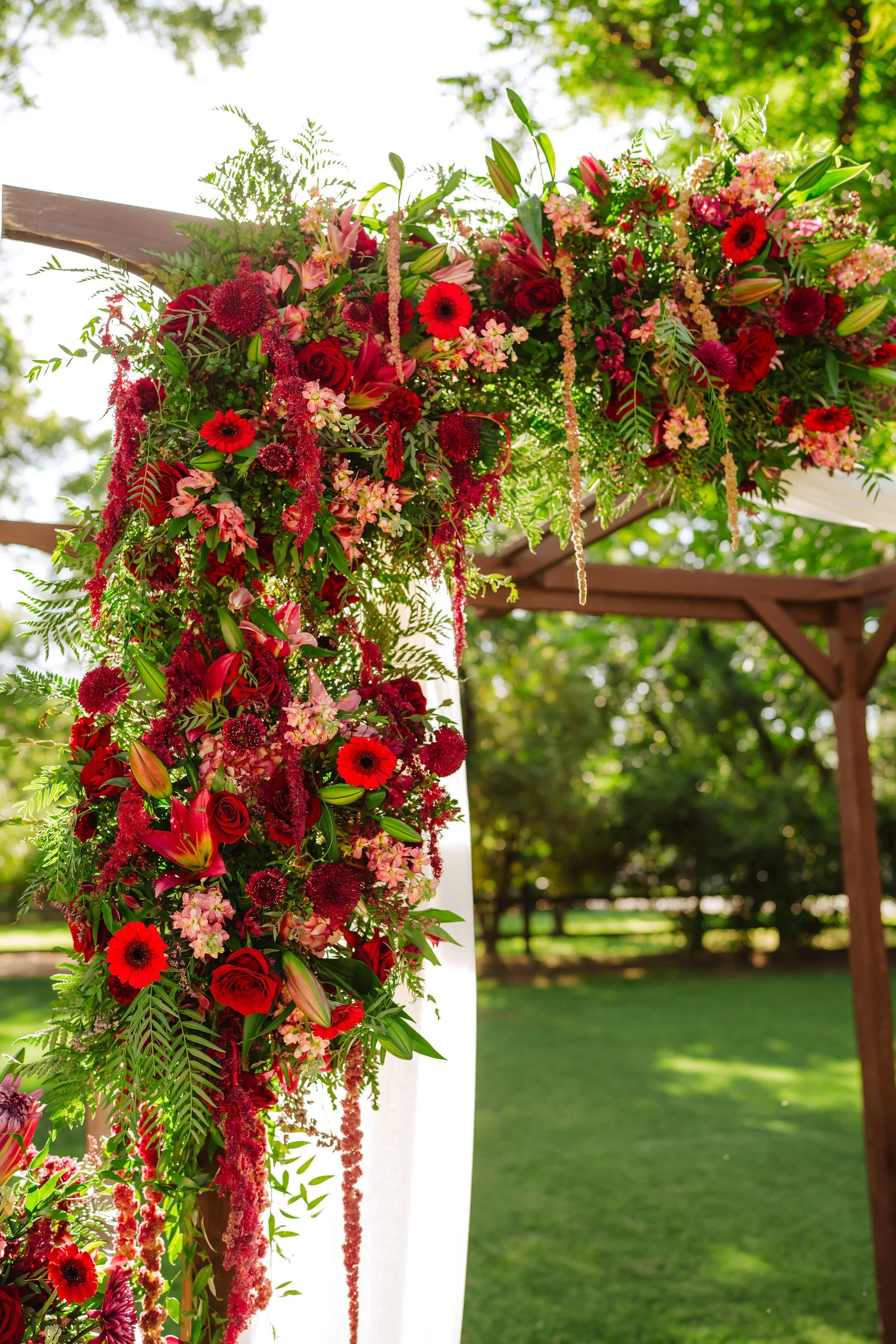 Statement wedding ceremony installation covered in cascading red, burgundy and pink flowers with organic greenery.