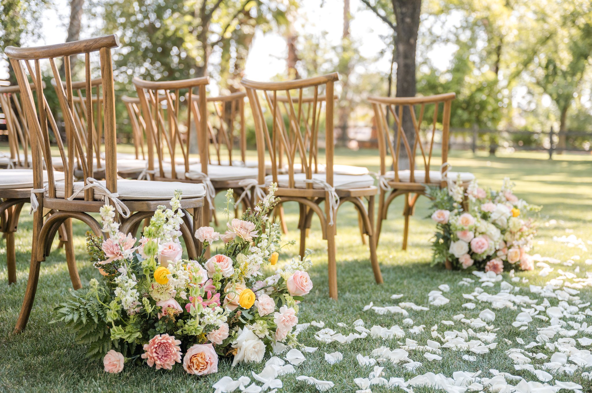 Colorful wedding aisle flowers with peach, pink and yellow blooms surrounding wooden ceremony chairs.