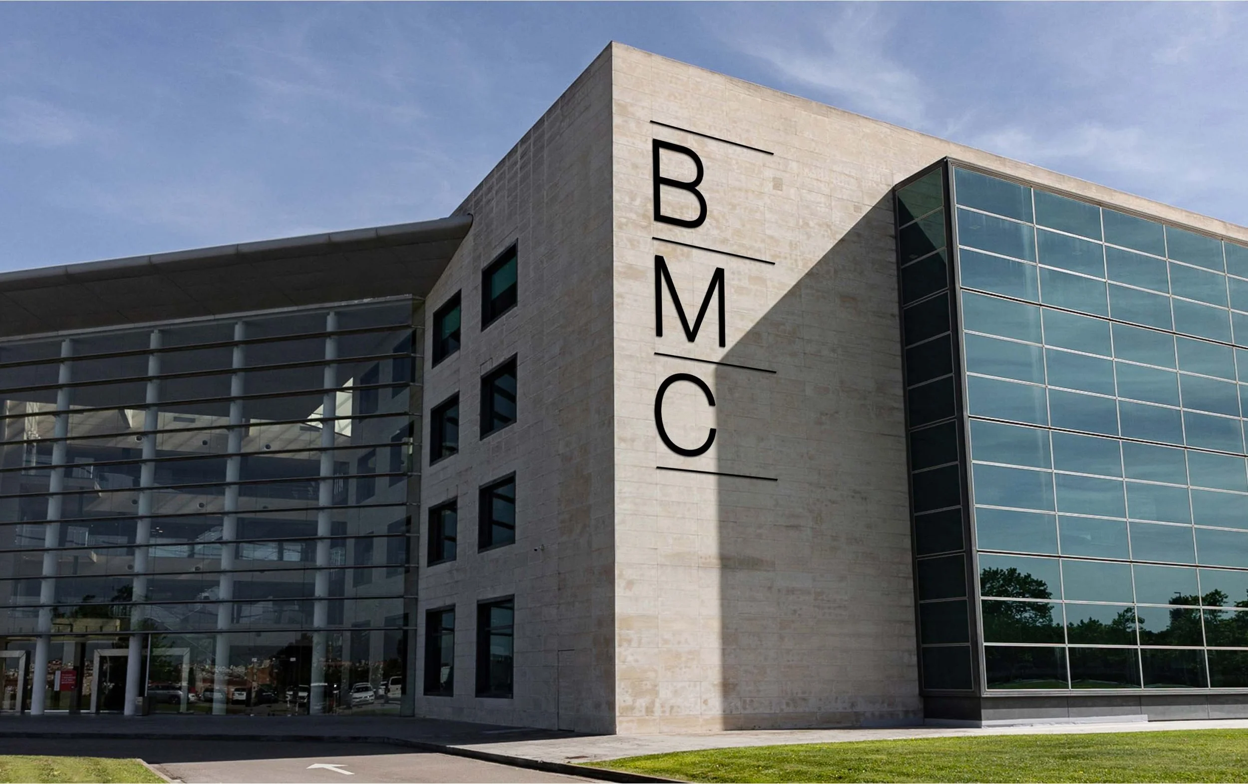 Edificio moderno con letras B, M, C en la esquina. Fachada de cristal y piedra, césped y camino en primer plano.