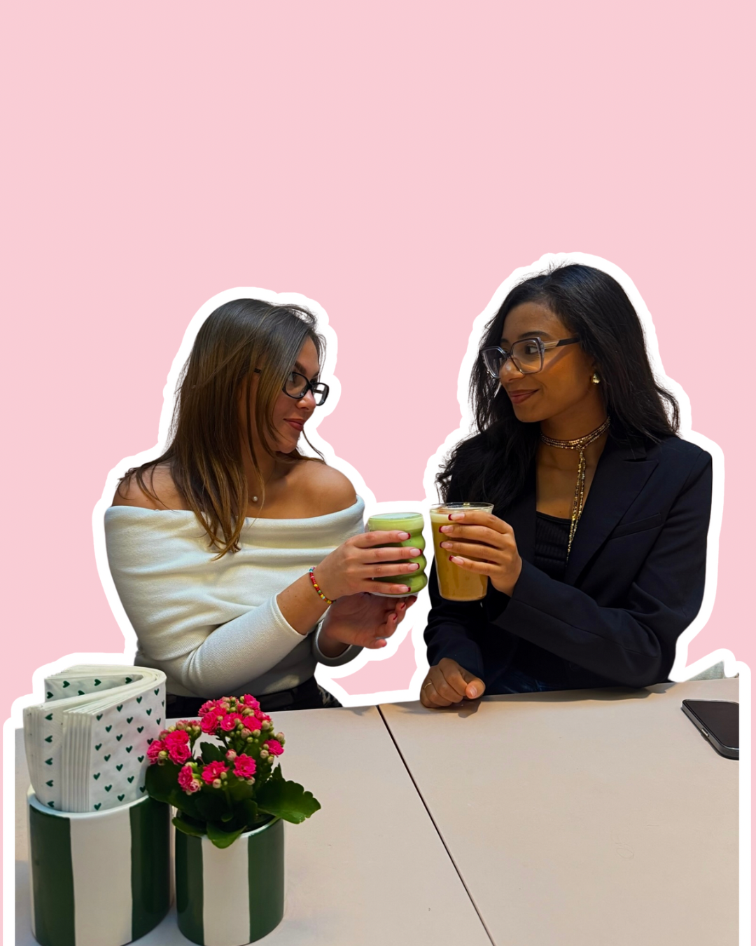 Deux femmes portant des lunettes, se tenant et levant des verres avec des boissons, avec un fond rose clair, une petite plante en pot et un napperon en papier avec un motif de cœurs sur la table.