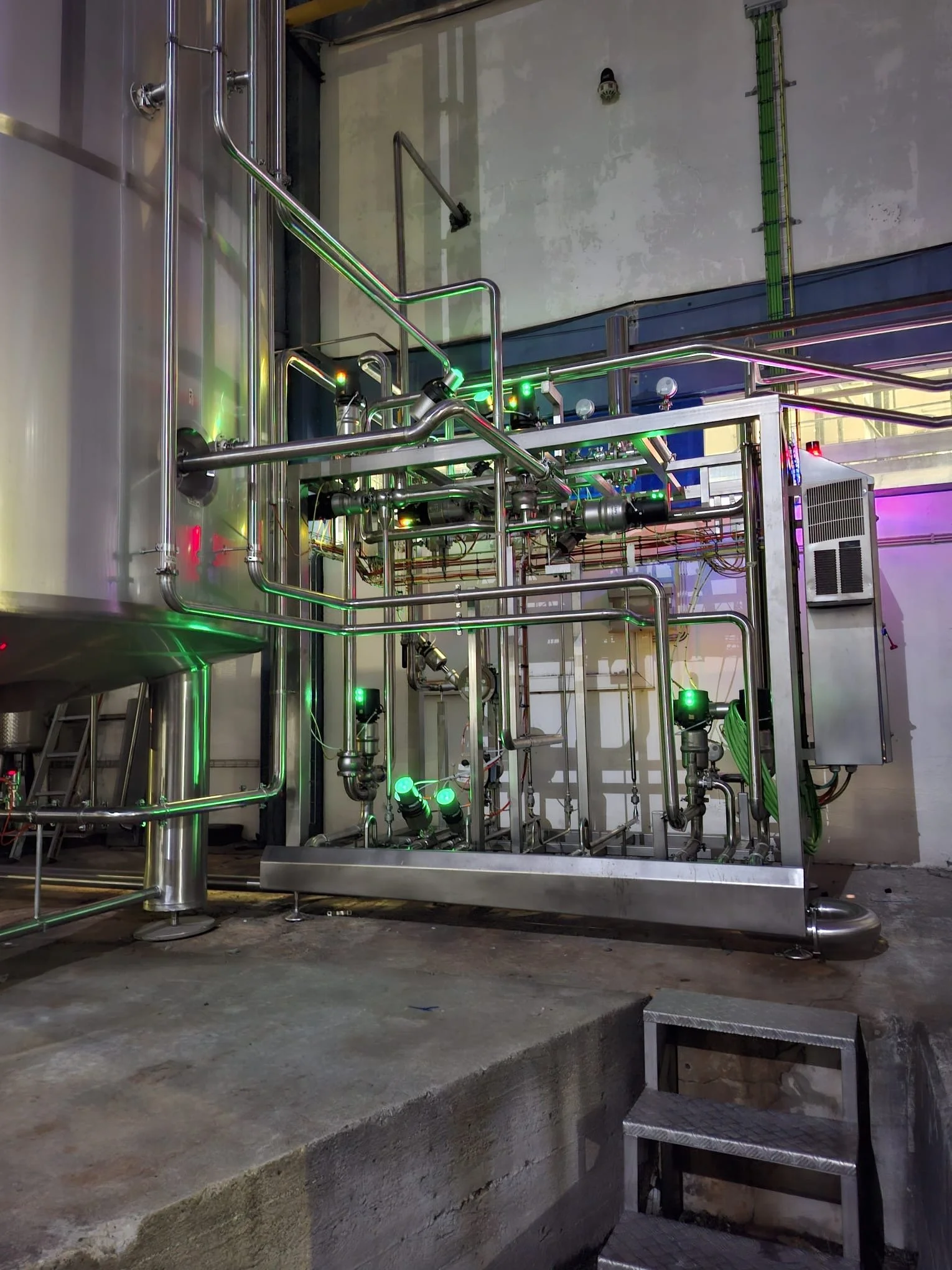 Industrial stainless-steel piping system with green and purple LED lights in a factory or processing plant.
