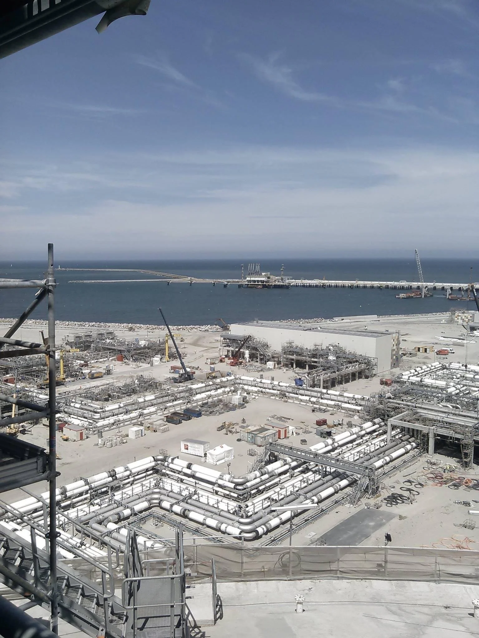 View of an industrial construction site near a body of water, with pipelines, equipment, and scaffolding, and a pier in the background under a partly cloudy sky.